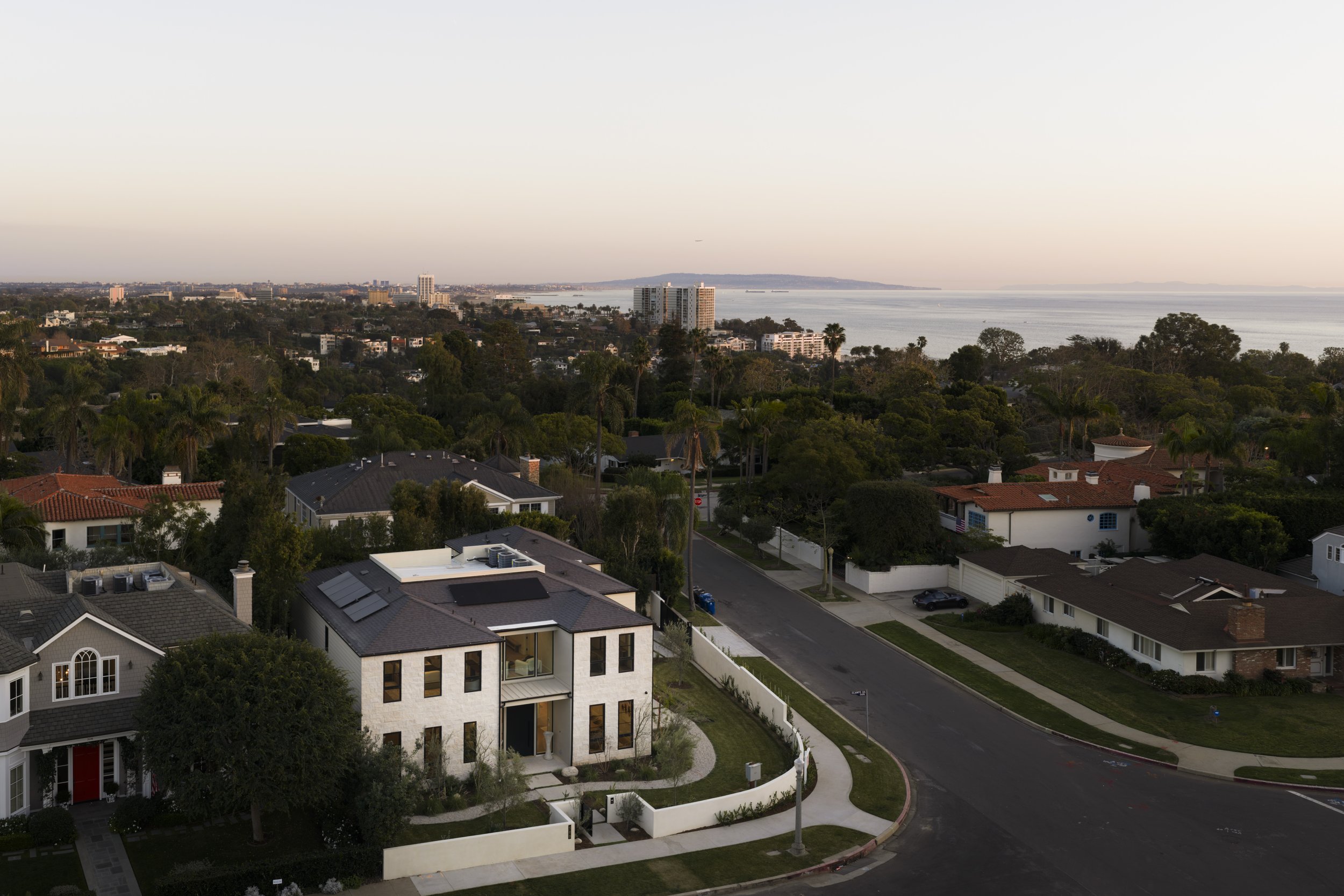 Residential neighborhood featuring modern white house in foreground, surrounded by trees and other homes, with city skyline and ocean in the background during sunset.