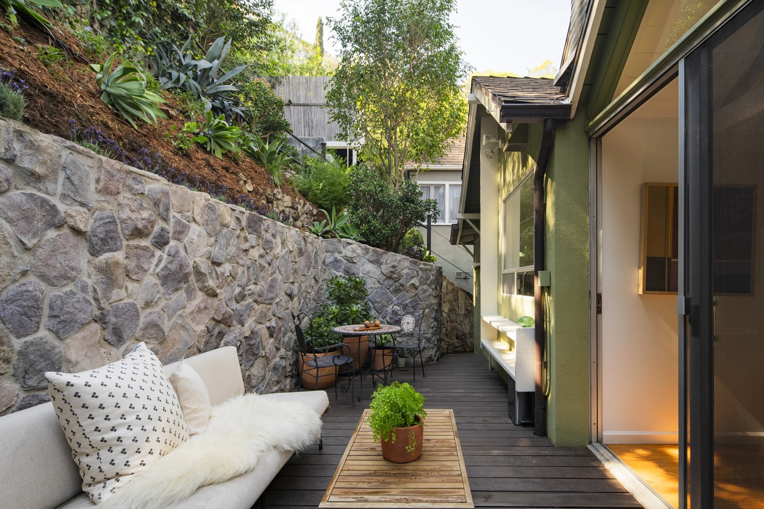 Outdoor patio area with wooden deck, stone wall, green plants, and outdoor furniture including a white sofa with pillows, a wooden coffee table with a potted fern, and a small black metal table with chairs.