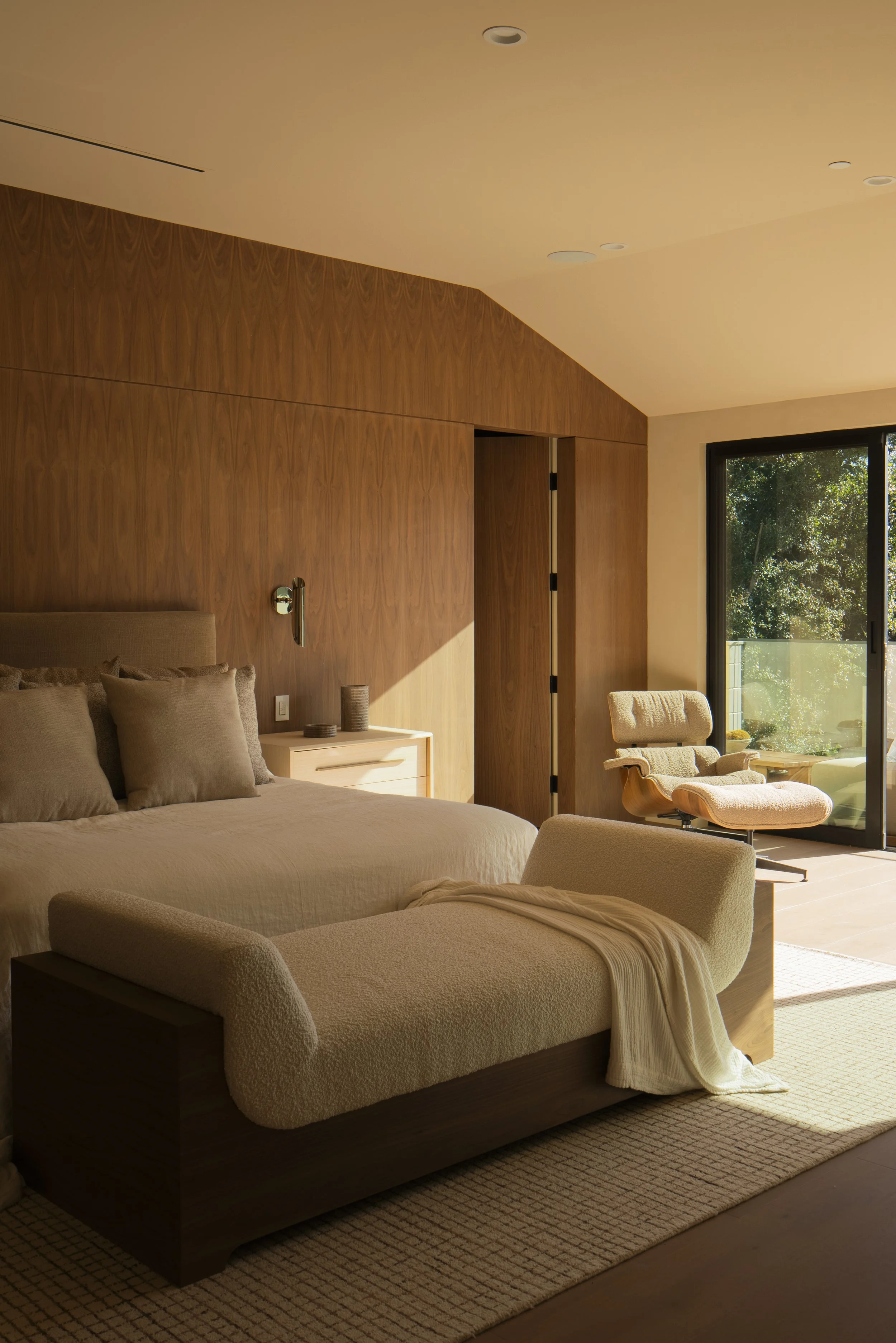 Modern bedroom with large bed, beige bedding, wooden wall, and a sitting chair near a sliding glass door.