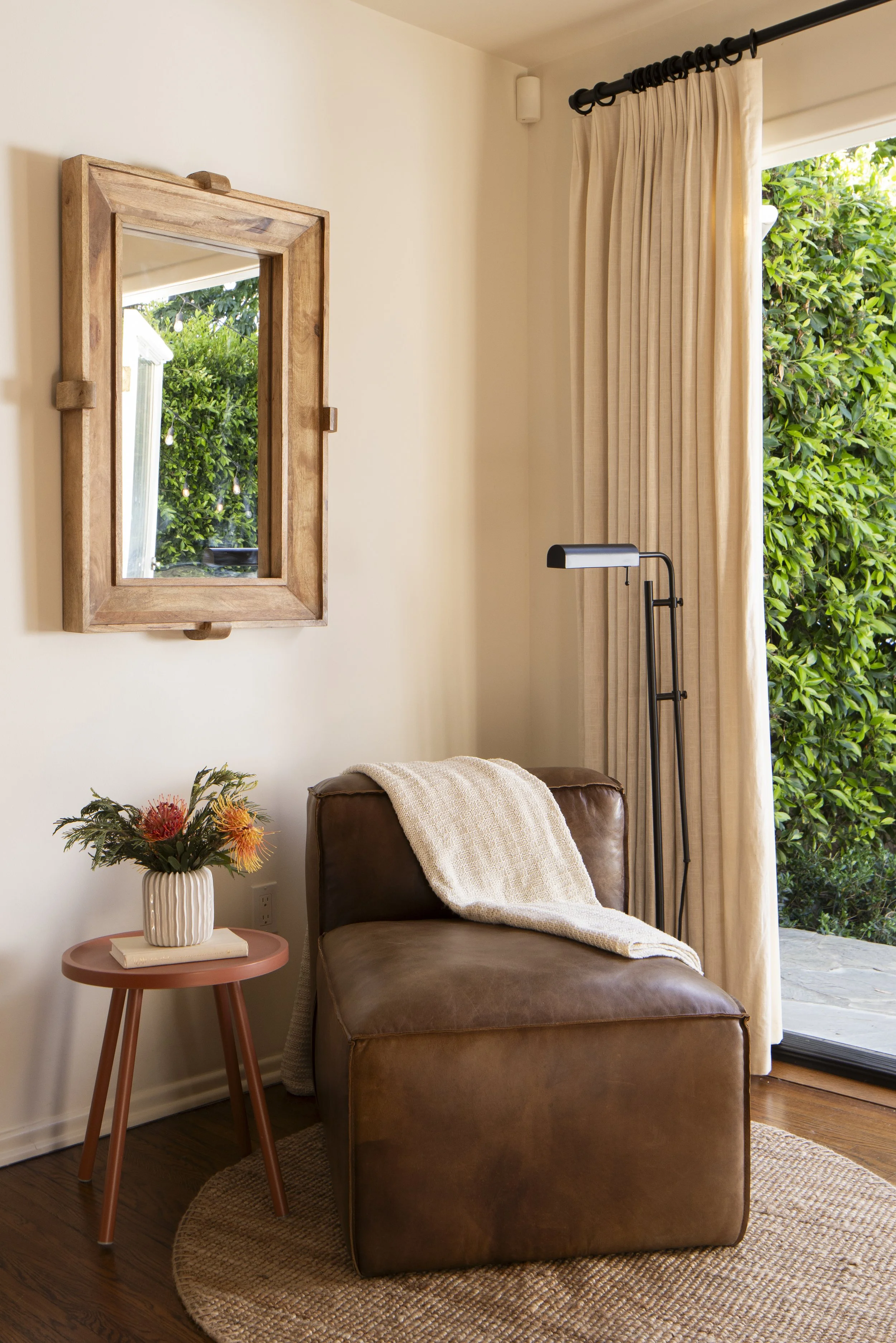 A cozy corner with a brown leather armchair, a white blanket draped over it, next to a small pink side table with a flower vase, a mirror on the wall, a floor lamp, beige curtains, and a sliding door with greenery outside.