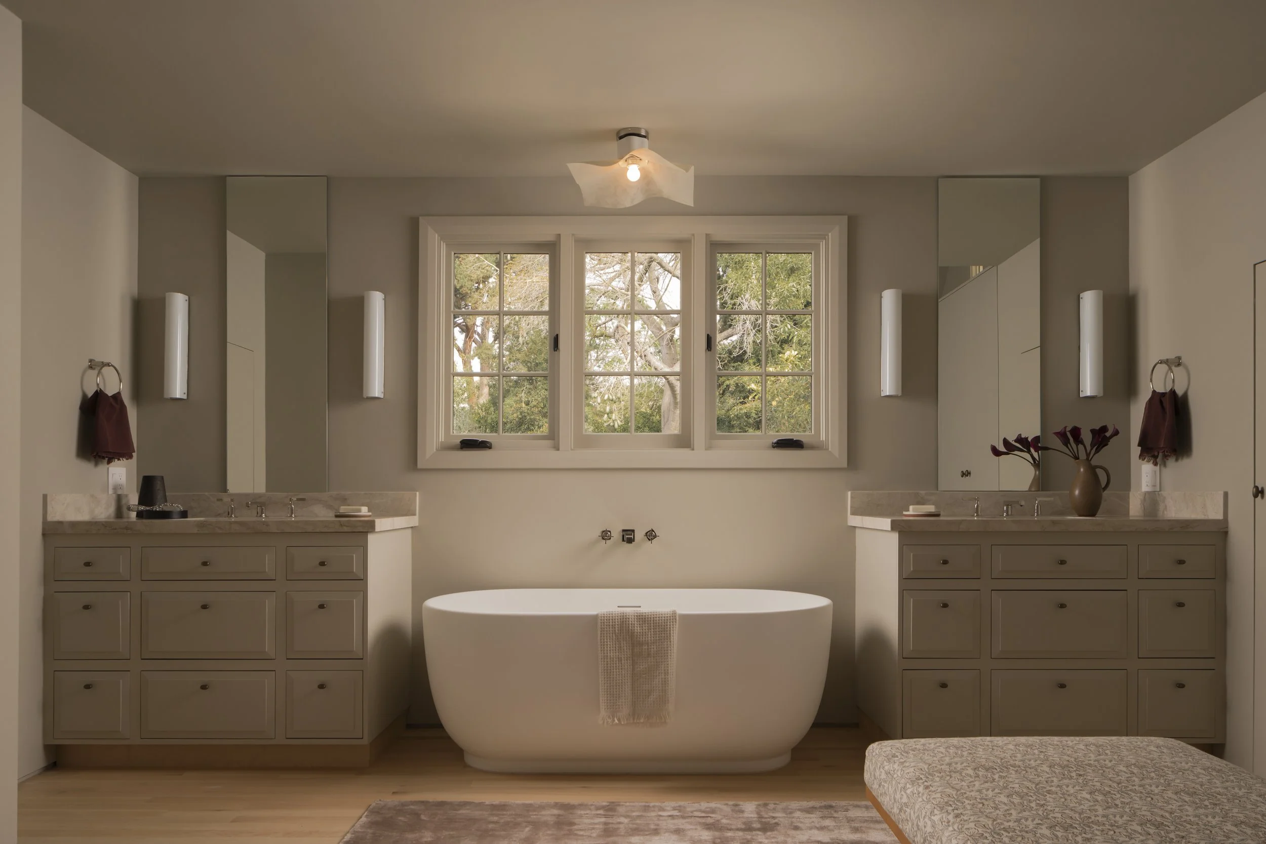 Bathroom with a freestanding bathtub centered below a window, with two vanity counters and mirrors on either side, and a beige rug at the foot of the tub.