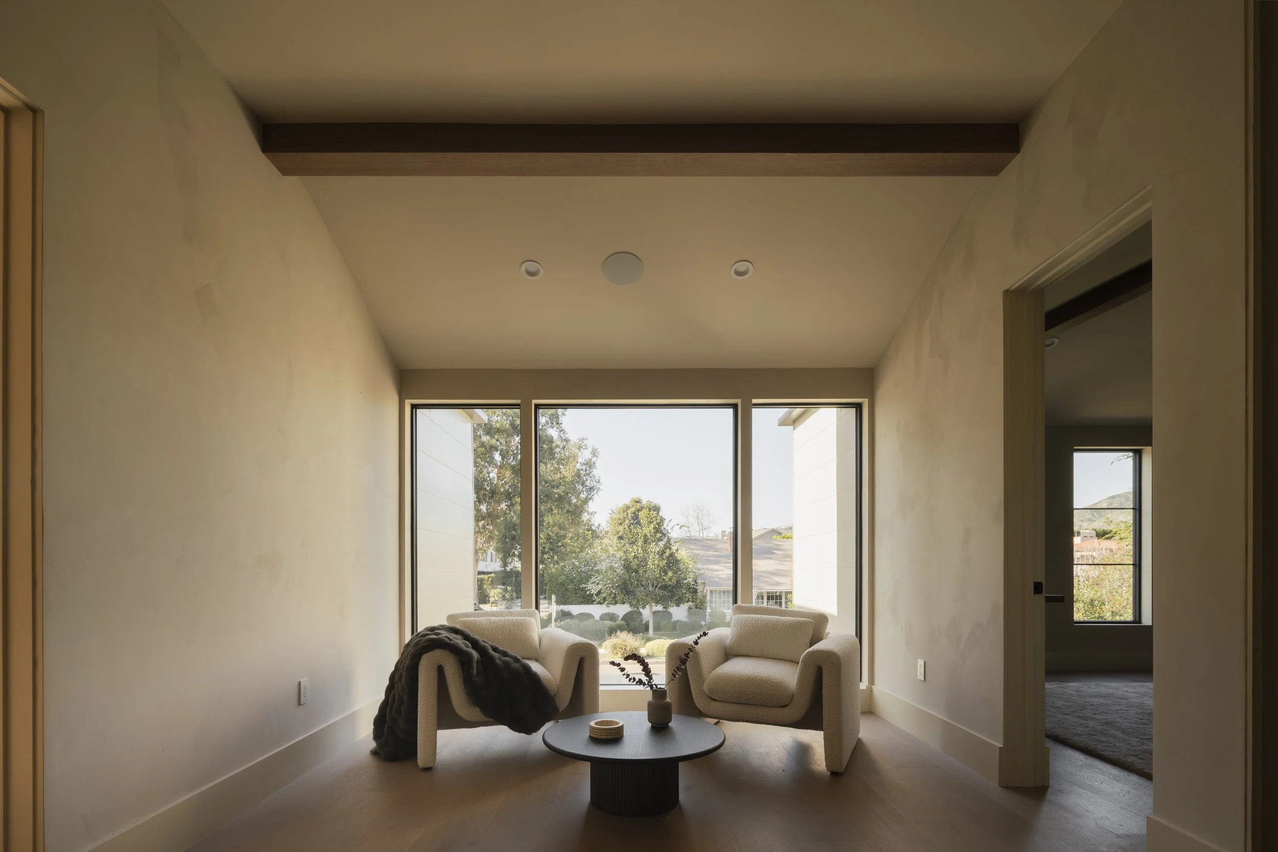 Living room with two beige armchairs, a black round coffee table with a vase and small dishes, large window revealing trees and houses outside, neutral wall colors, and wooden flooring.