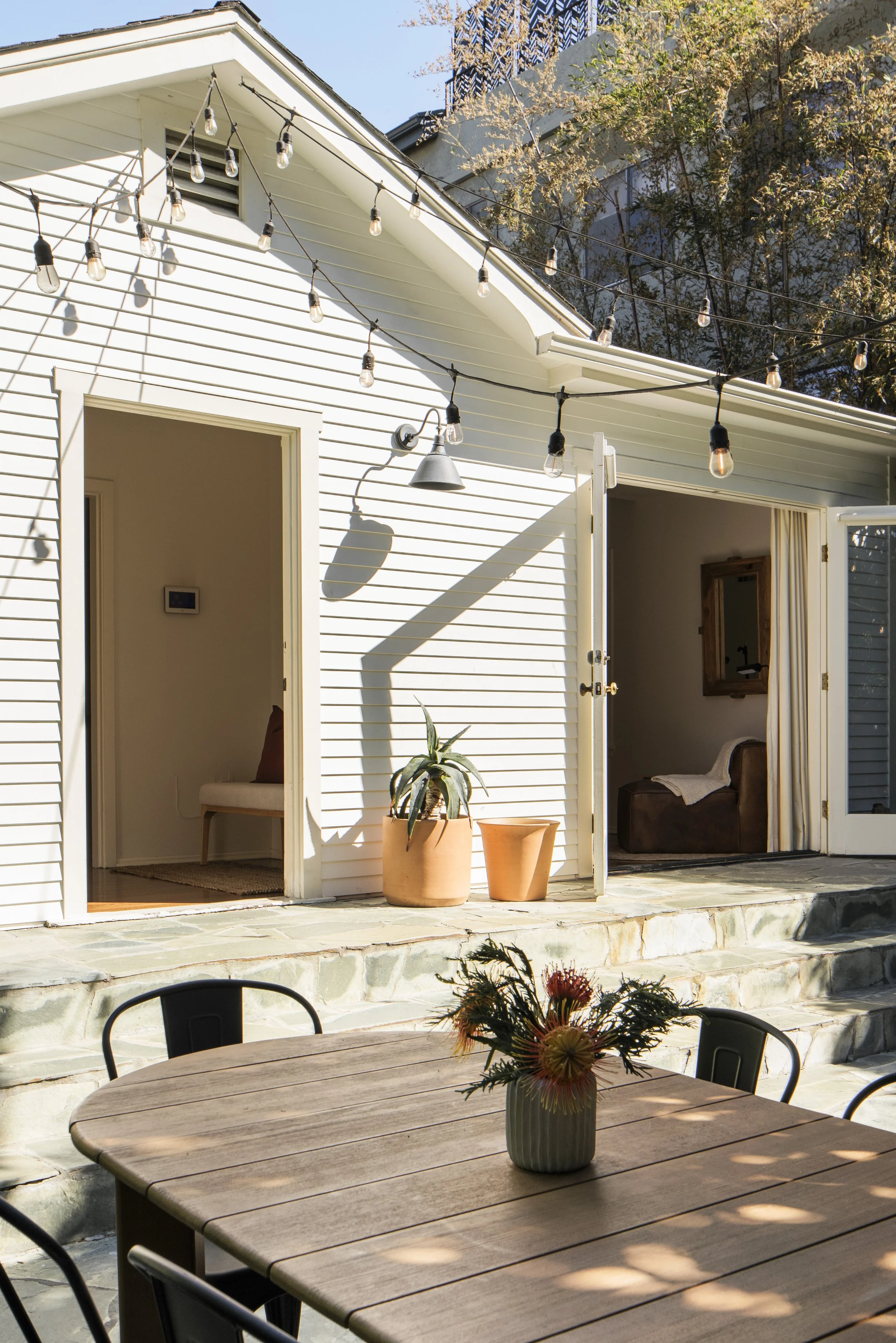 Outdoor patio with a wooden table and black chairs, potted plants, string lights, and open doors leading inside a white house.