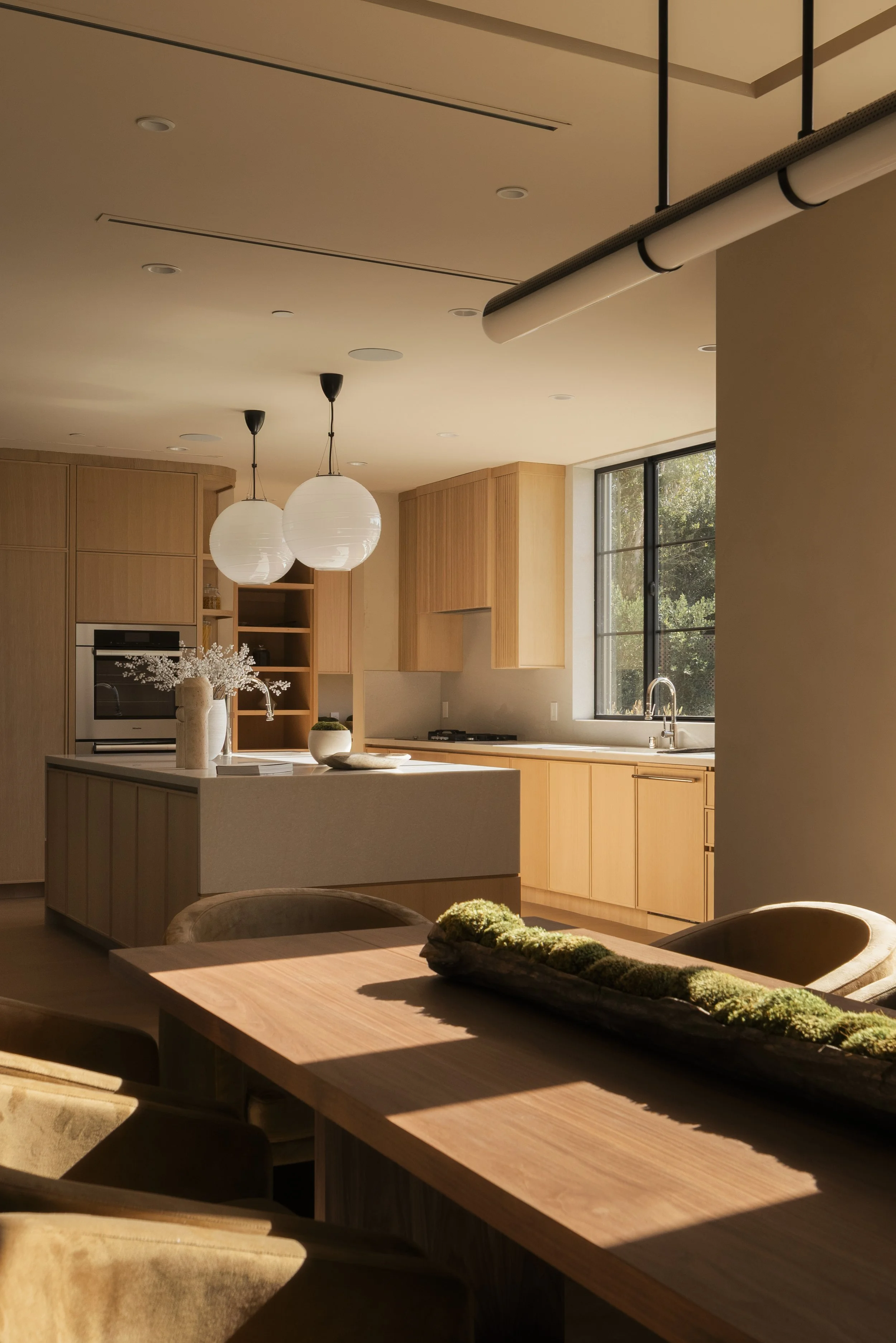 A modern kitchen and dining area with light wood cabinets, a white island, hanging pendant lights, a large window with black frames, and natural sunlight illuminating the space.