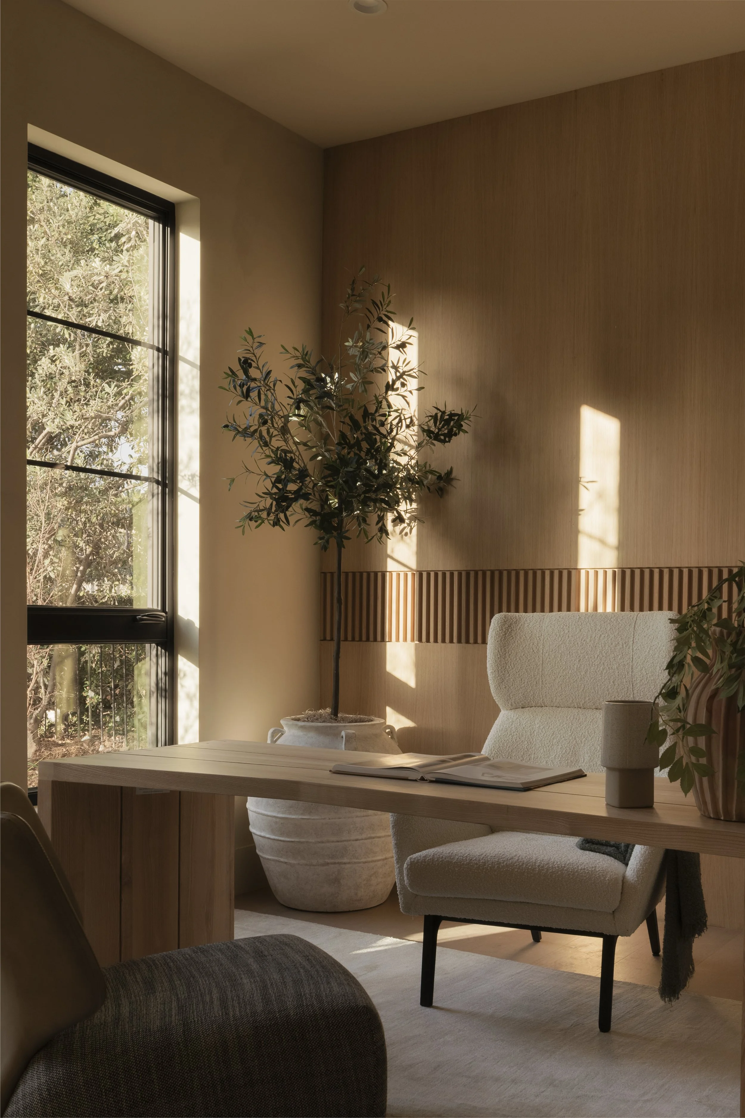 A cozy office corner with a large window, a white armchair, a wooden desk with an open book, a beige mug, a potted plant, and sunlight casting shadows on the wall.