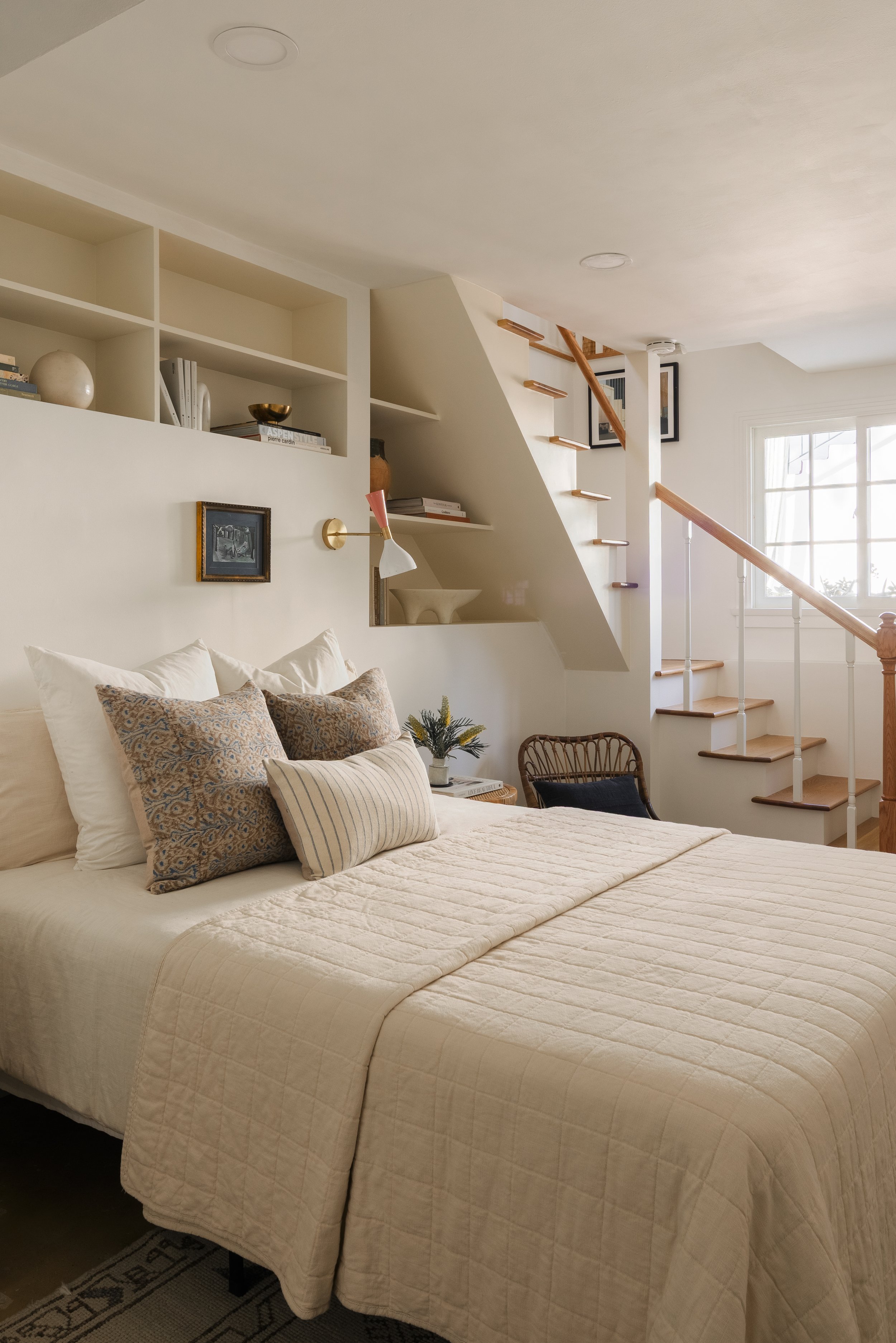 A cozy bedroom with a bed covered in a beige quilt, decorated with patterned and striped pillows, next to a small bedside table with a plant. In the background, there is a staircase with wooden steps and a white railing, and a window letting in natur
