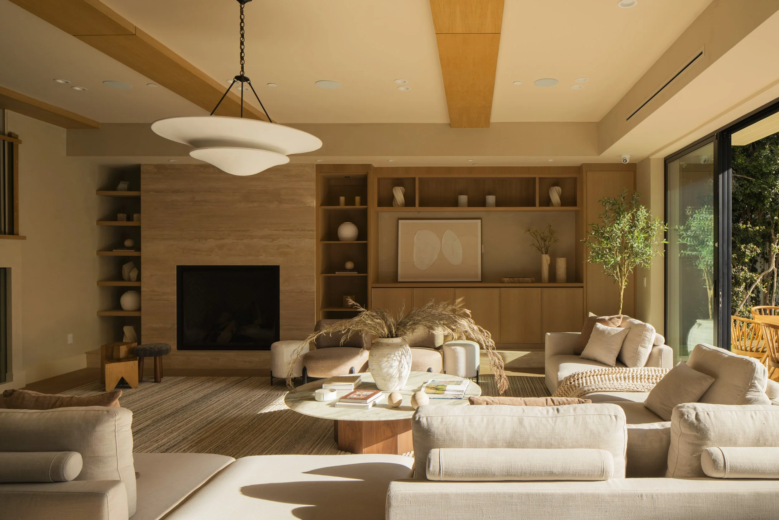 A living room with a beige sofa set, a round white coffee table, and a wall-mounted flat-screen TV. There is a modern chandelier above, built-in wooden shelves with decorative items, and large sliding glass doors opening to an outdoor patio with a wo