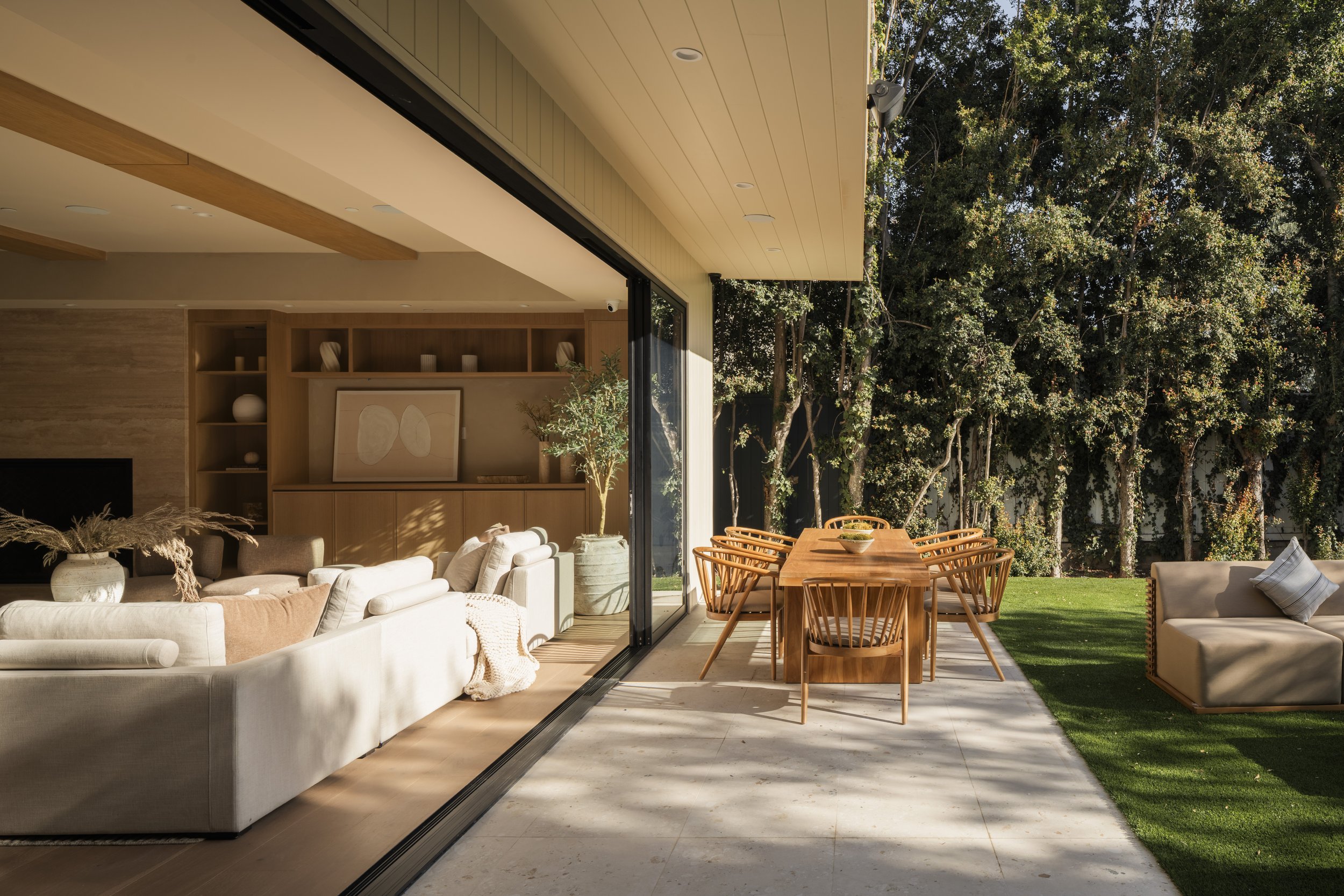 Living room with beige sofas and artwork, open to an outdoor patio with a wooden dining table and chairs, surrounded by green trees and grass.