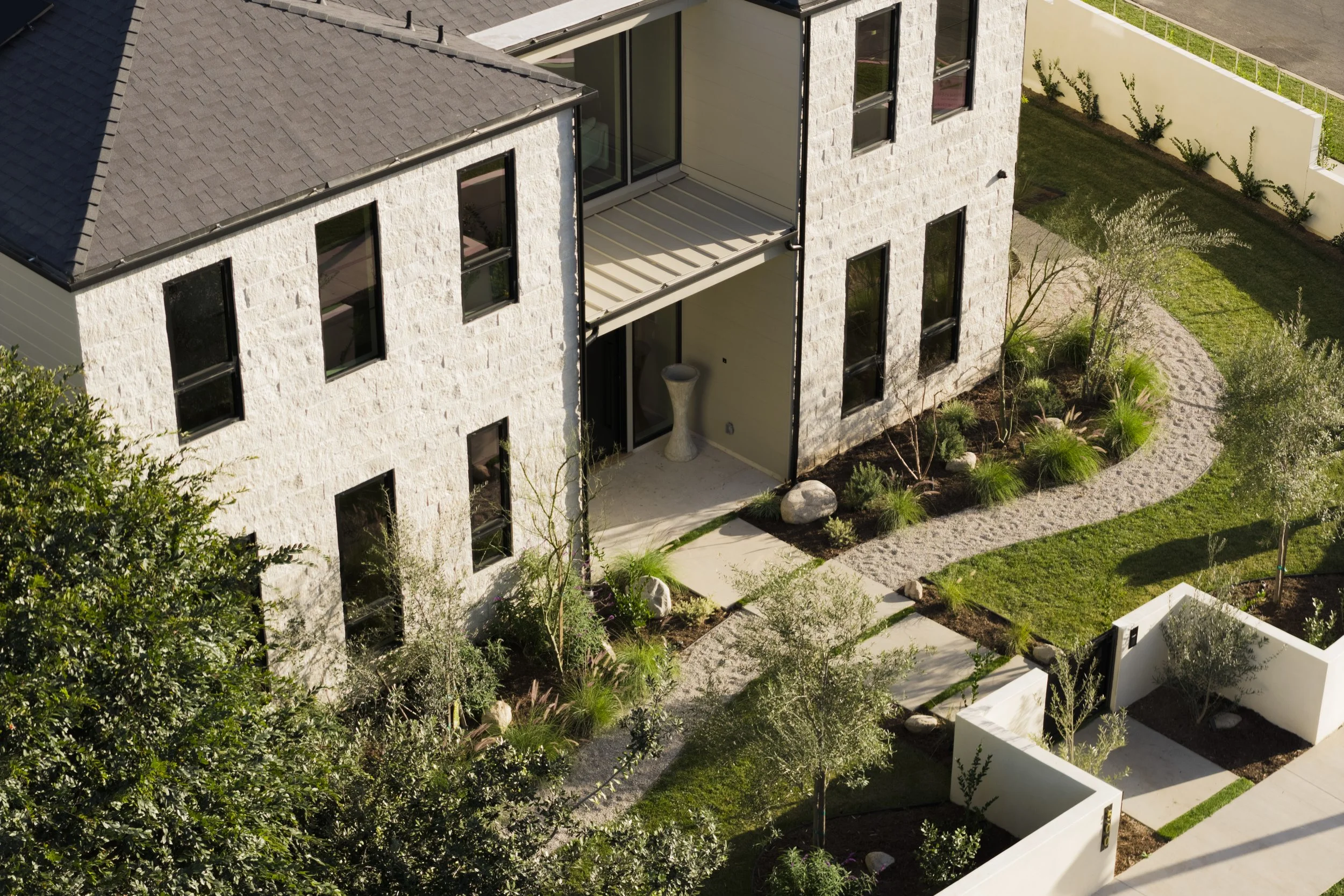 Aerial view of a modern, multi-story residential building with light-colored stone exterior, multiple black-framed windows, and a small balcony. Surrounding the building are landscaped lawns with pathways, small trees, and shrubs.