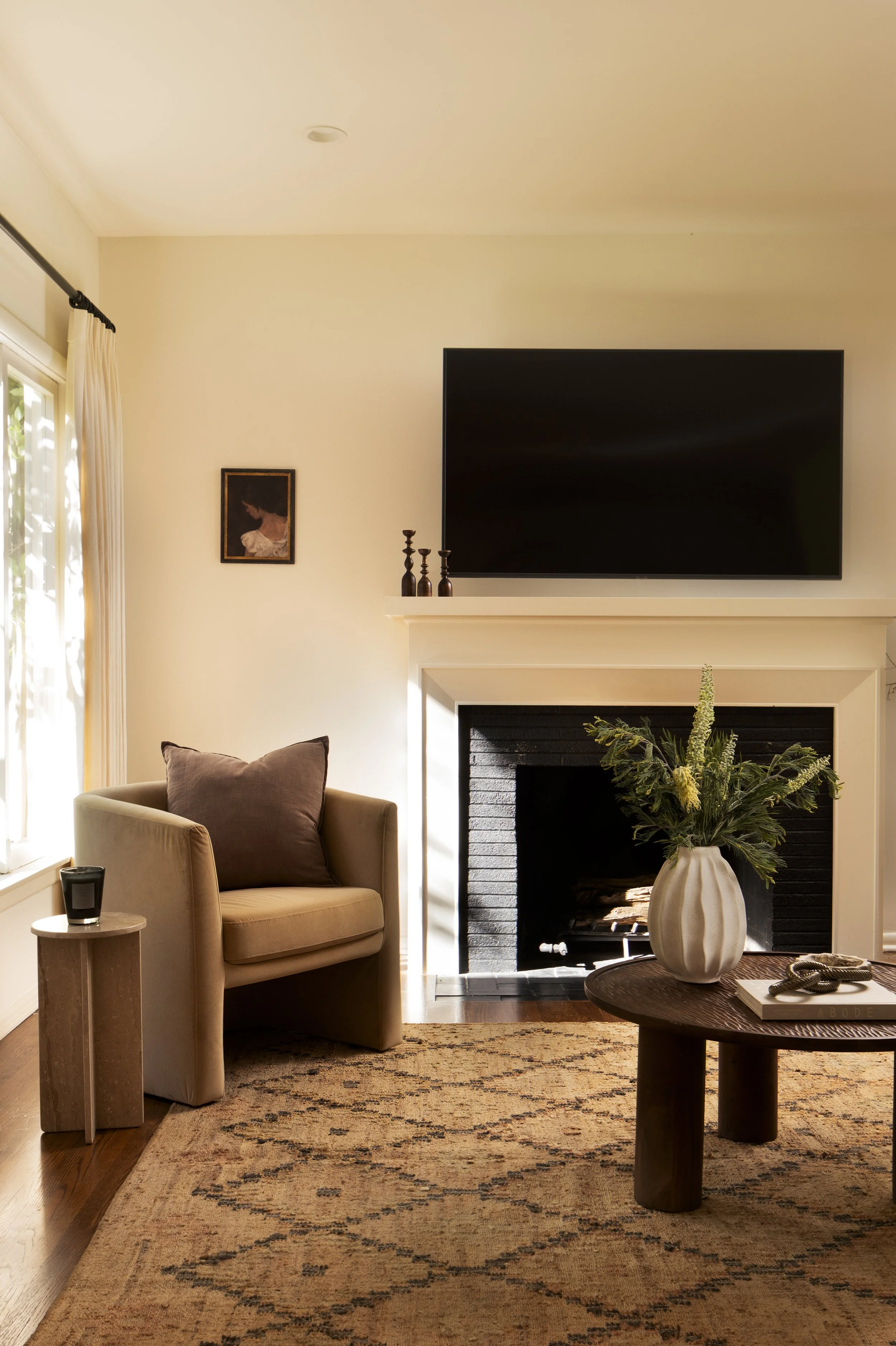 Living room with a fireplace, wall-mounted TV, beige armchair with large cushion, side table with a speaker, a round wooden coffee table with a large vase of flowers, framed artwork on the wall, and a patterned rug.