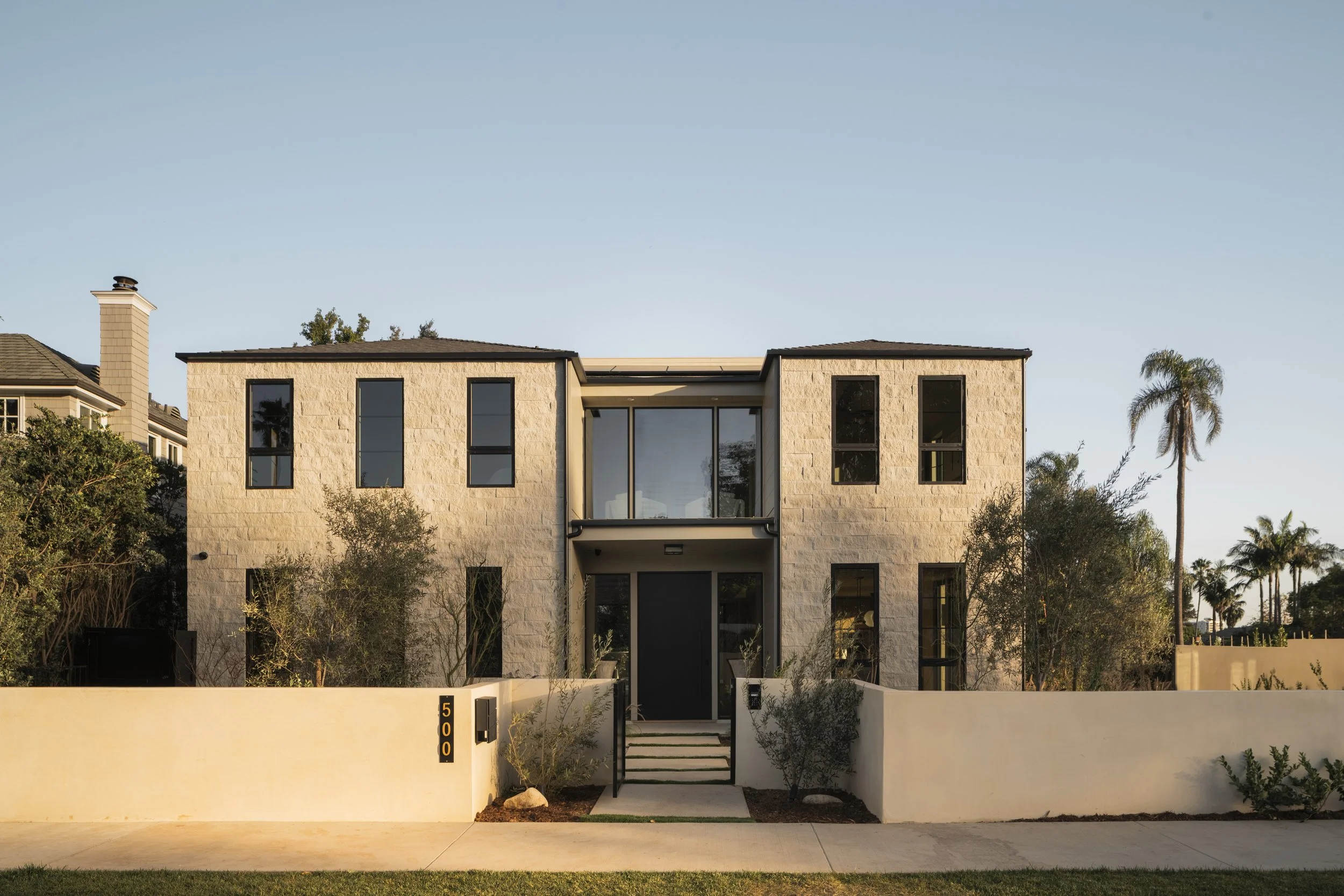 Modern two-story house with tan stone exterior, black window frames, front steps, and a white wall at the sidewalk.