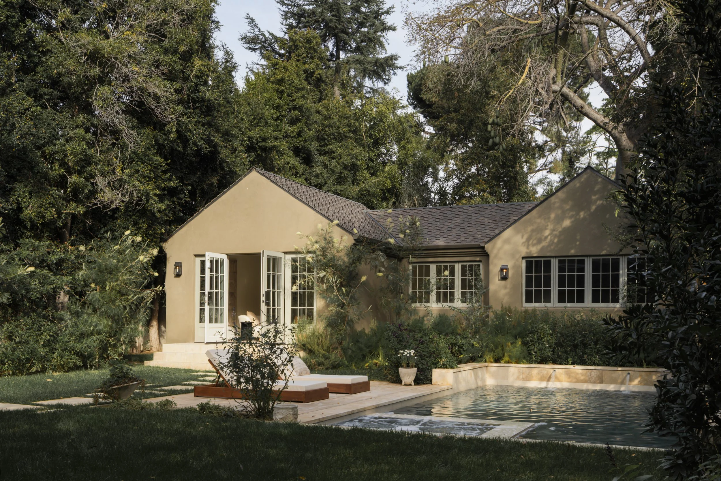 A house with beige walls, multiple windows, and open French doors, surrounded by lush trees and greenery. There is a swimming pool in the foreground with a poolside lounge chair and potted plants nearby.