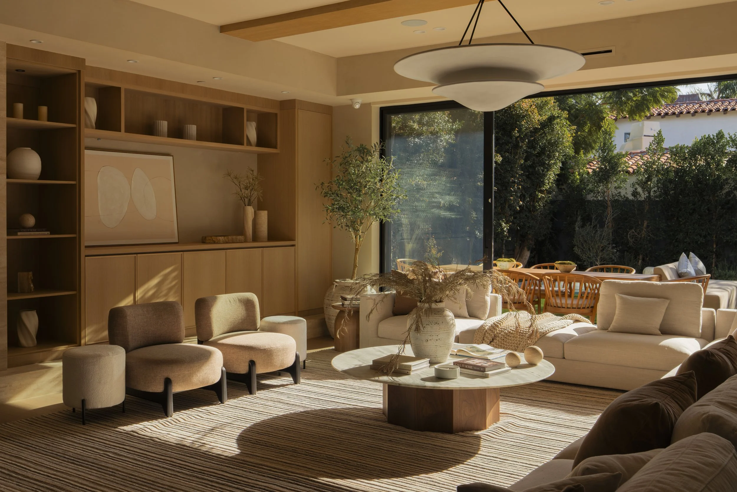 Living room with a large window, beige and white sofas, a round coffee table with vases and books, and wooden shelving and dining area in the background