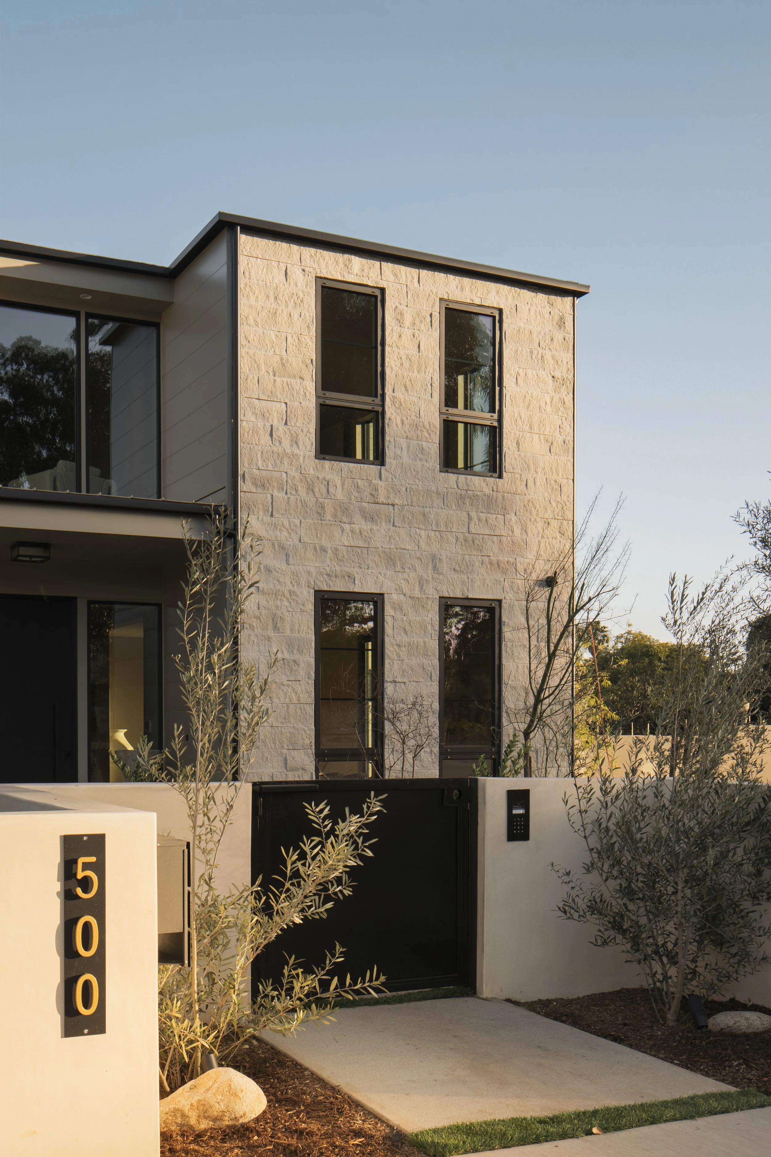 Modern multi-story house with beige stone exterior, black window frames, black gate, and minimalist landscaping including small trees and rocks, during daytime with clear sky.