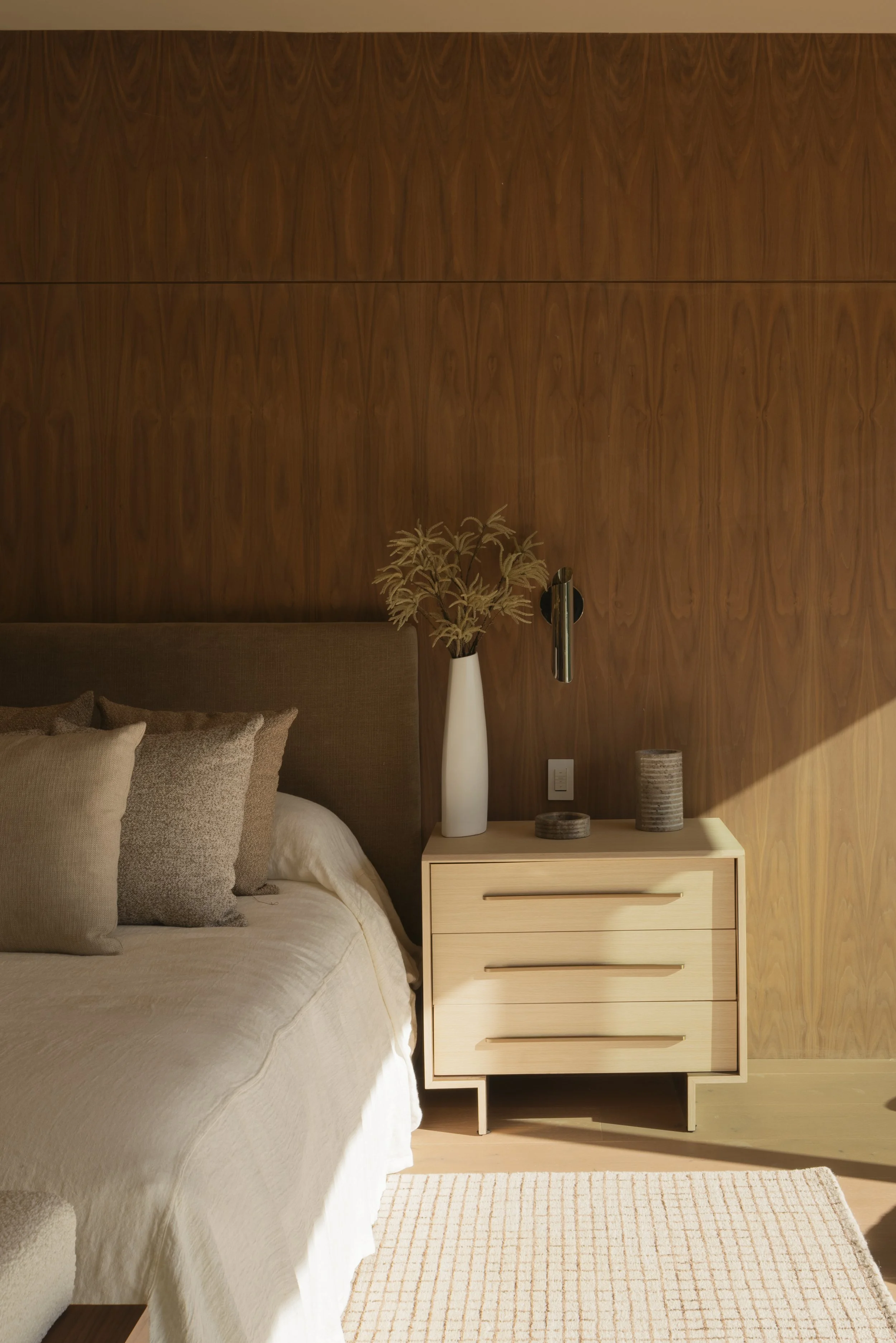 A bedroom with a bed, beige bedding, and pillows, a wooden nightstand with a white vase containing decorative branches, and two small round containers on top, against a wood-paneled wall with sunlight coming in.
