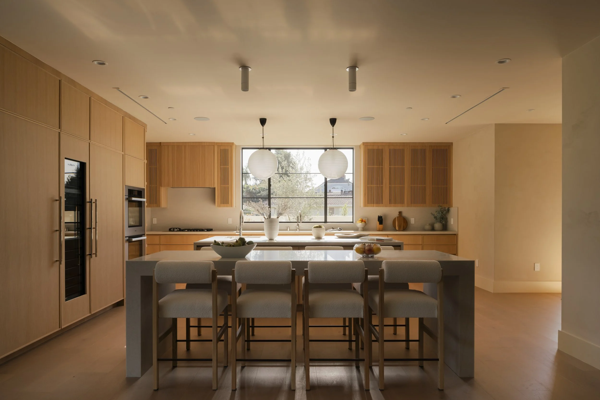 Modern kitchen with wooden cabinetry, large island with seating, and two pendant lights. Large window behind the sink, decorated with bowls and small plants.