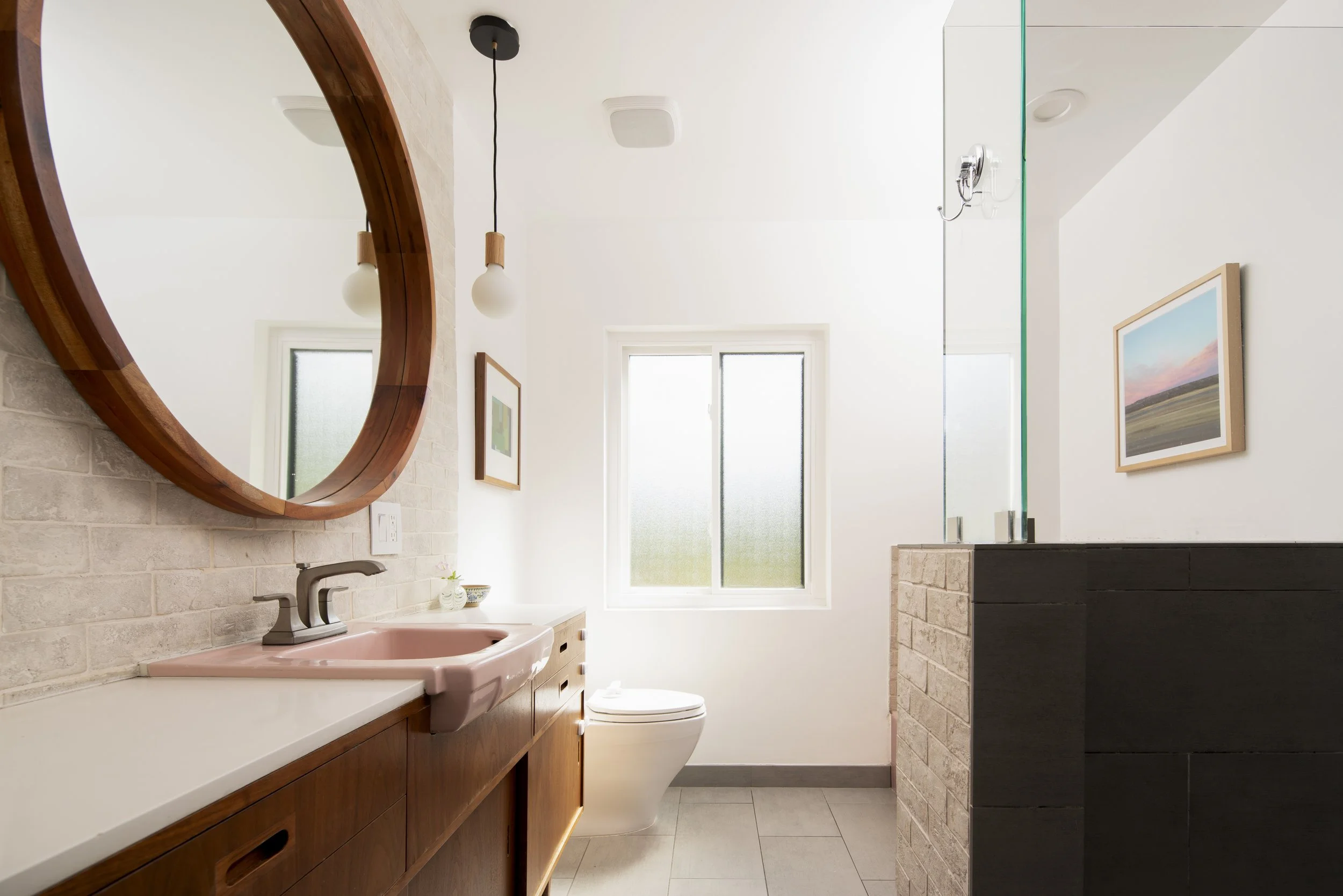 Modern bathroom with a pink sink, wooden vanity, round mirror, framed artwork, frosted window, and minimalist light fixtures.