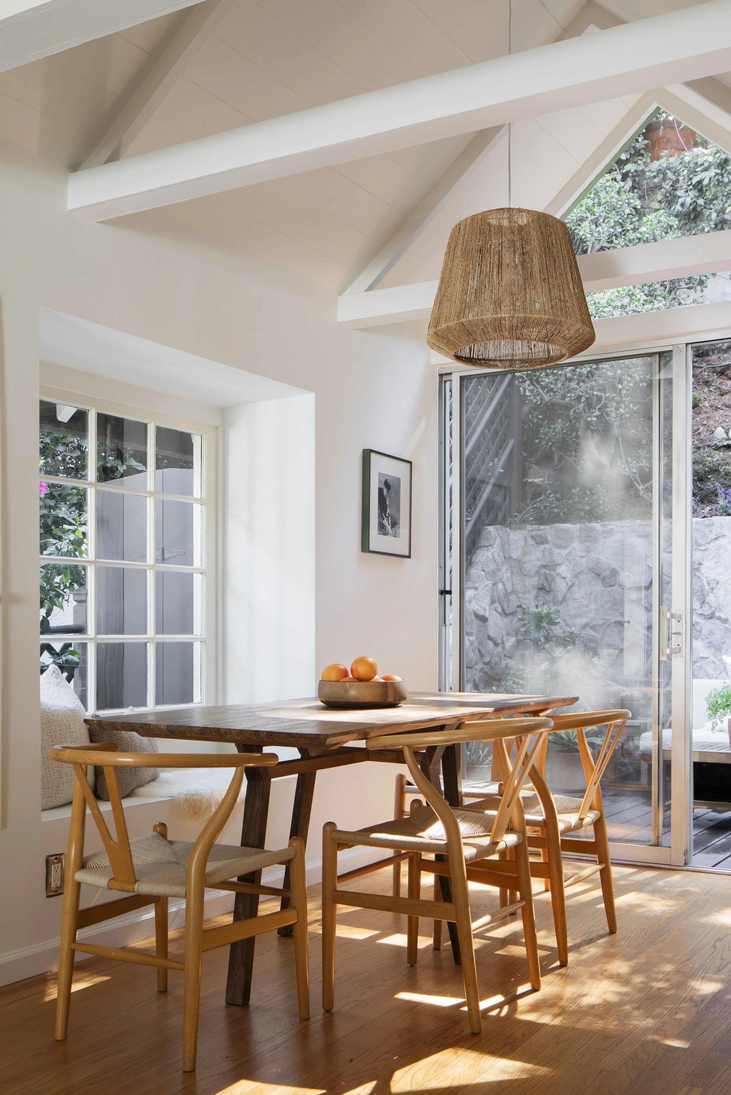 A sunlit dining area with a wooden table and four matching chairs, a bowl of oranges on the table, a window seat cushion, a black-and-white framed photograph on the wall, and a sliding glass door leading to an outdoor area with rocks and greenery. A 