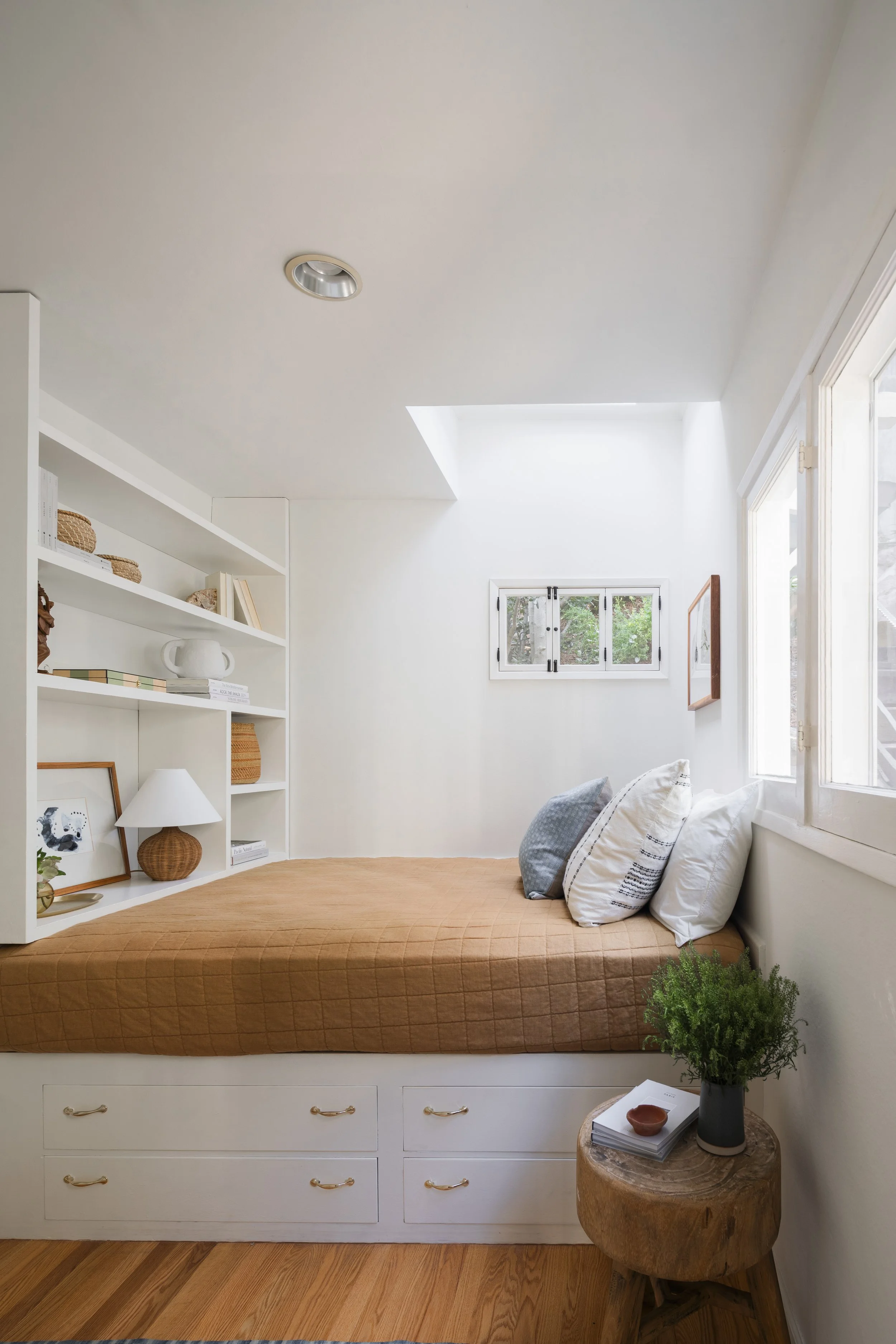 A cozy bedroom with a built-in bed featuring drawers underneath, an orange quilt, and three pillows. To the right, there is a small round wooden side table with a potted green plant, a small container, and a stack of books. The room has white walls, 