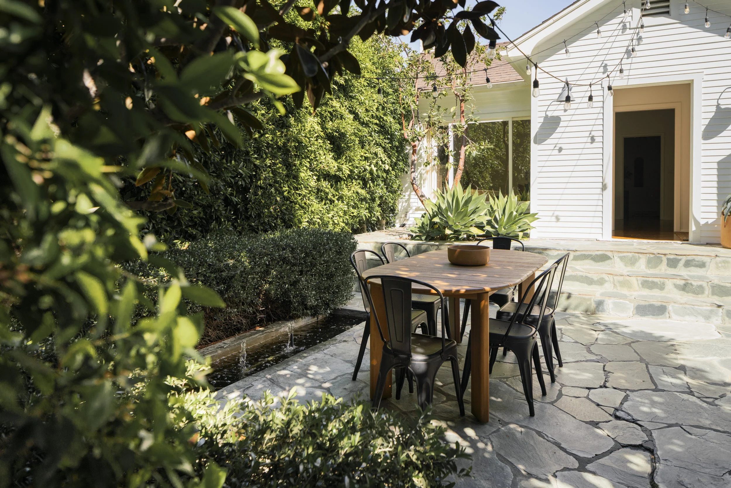 Outdoor patio with a wooden dining table, six black chairs, a pond with small water features, lush green bushes, large agave plants, and a white house with string lights hanging outside.