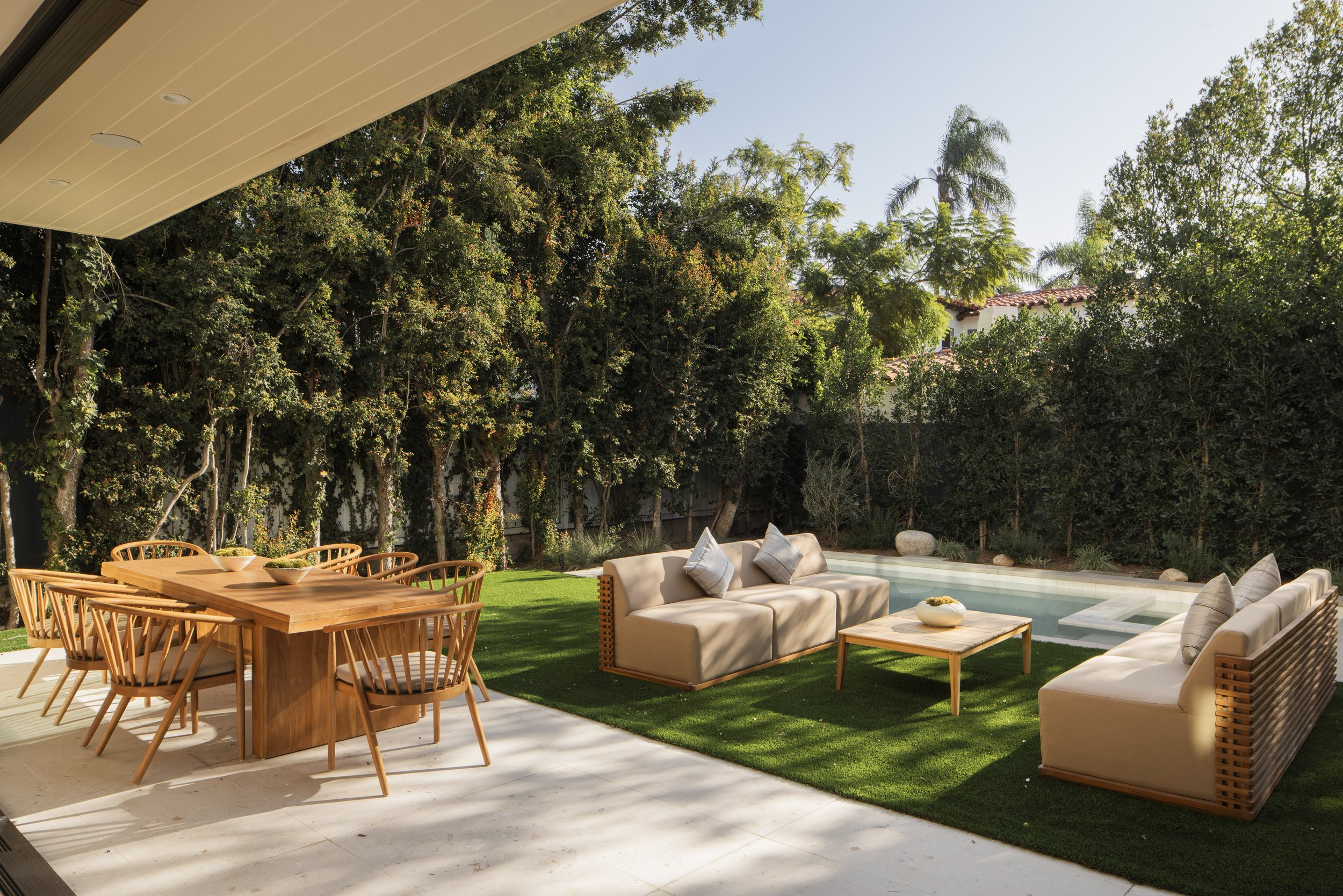 Outdoor patio with a wooden dining table and chairs, and cushioned sofas around a small wooden table with a pool in the background, surrounded by trees and greenery on a sunny day.