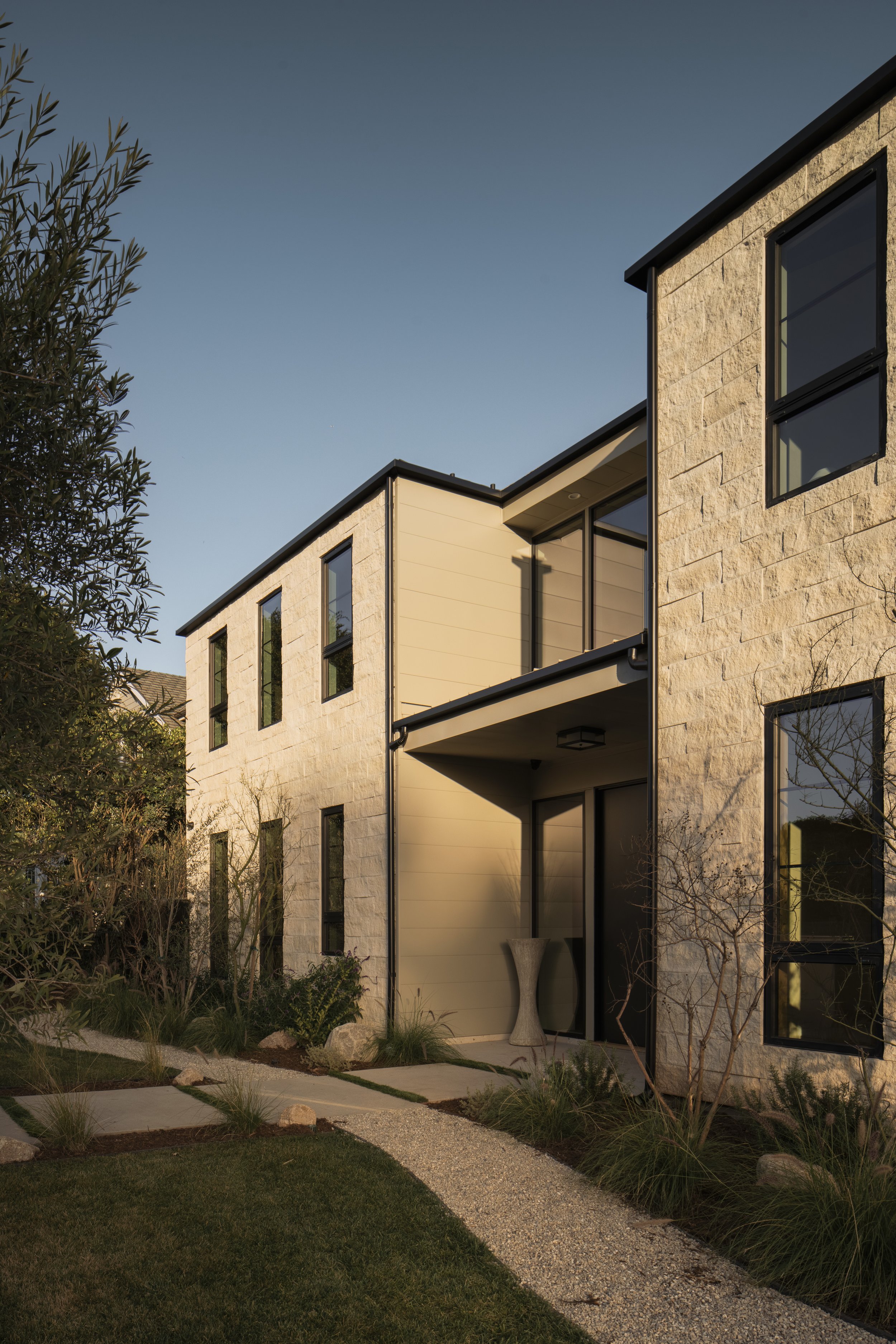 Modern residential building with beige stone walls, large windows, and a small garden with pathway at sunset.