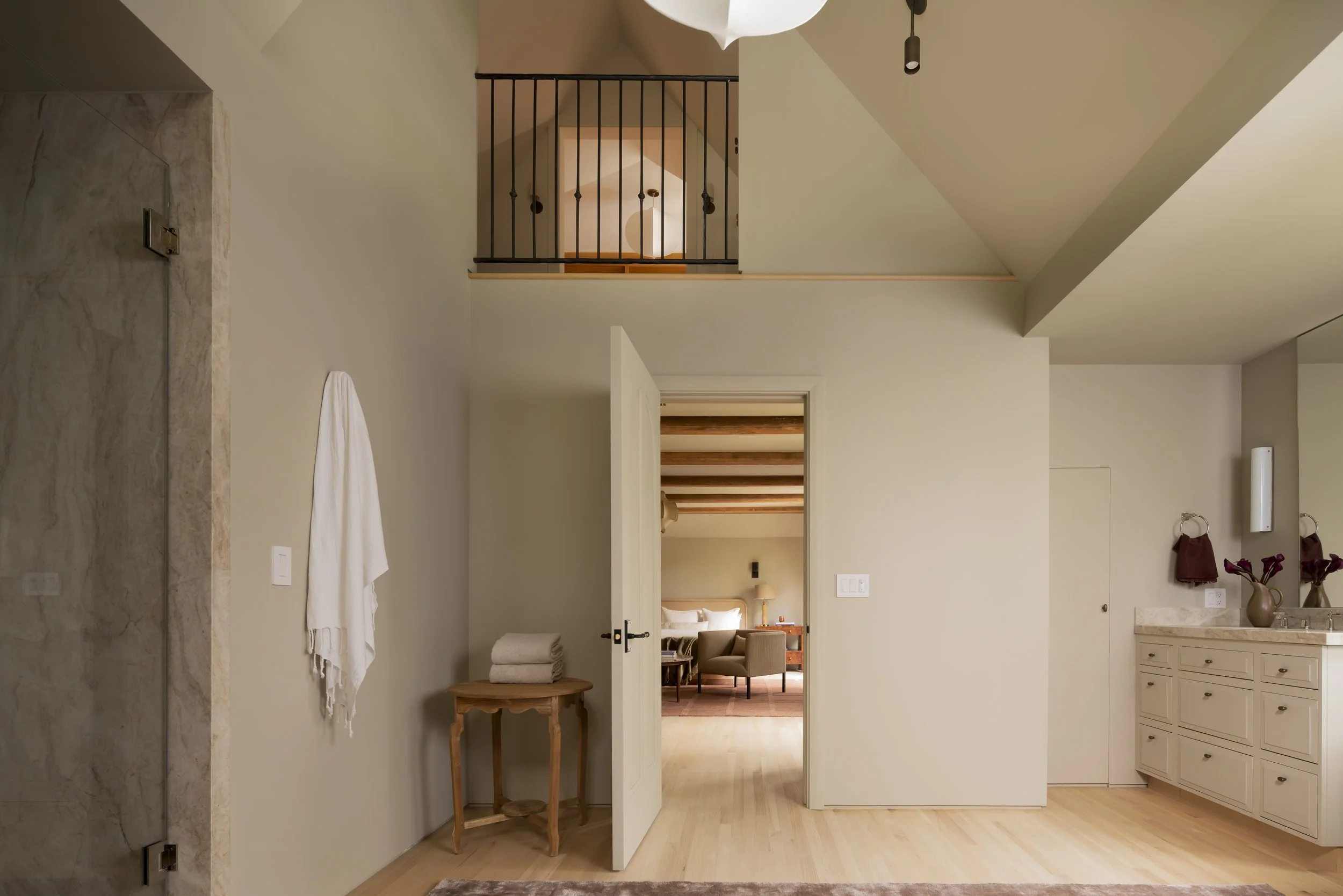 Interior view of a modern home featuring a doorway leading to a bedroom, a small wooden table with folded towels, a towel hanging on the wall, and a bathroom vanity with a mirror and flowers.