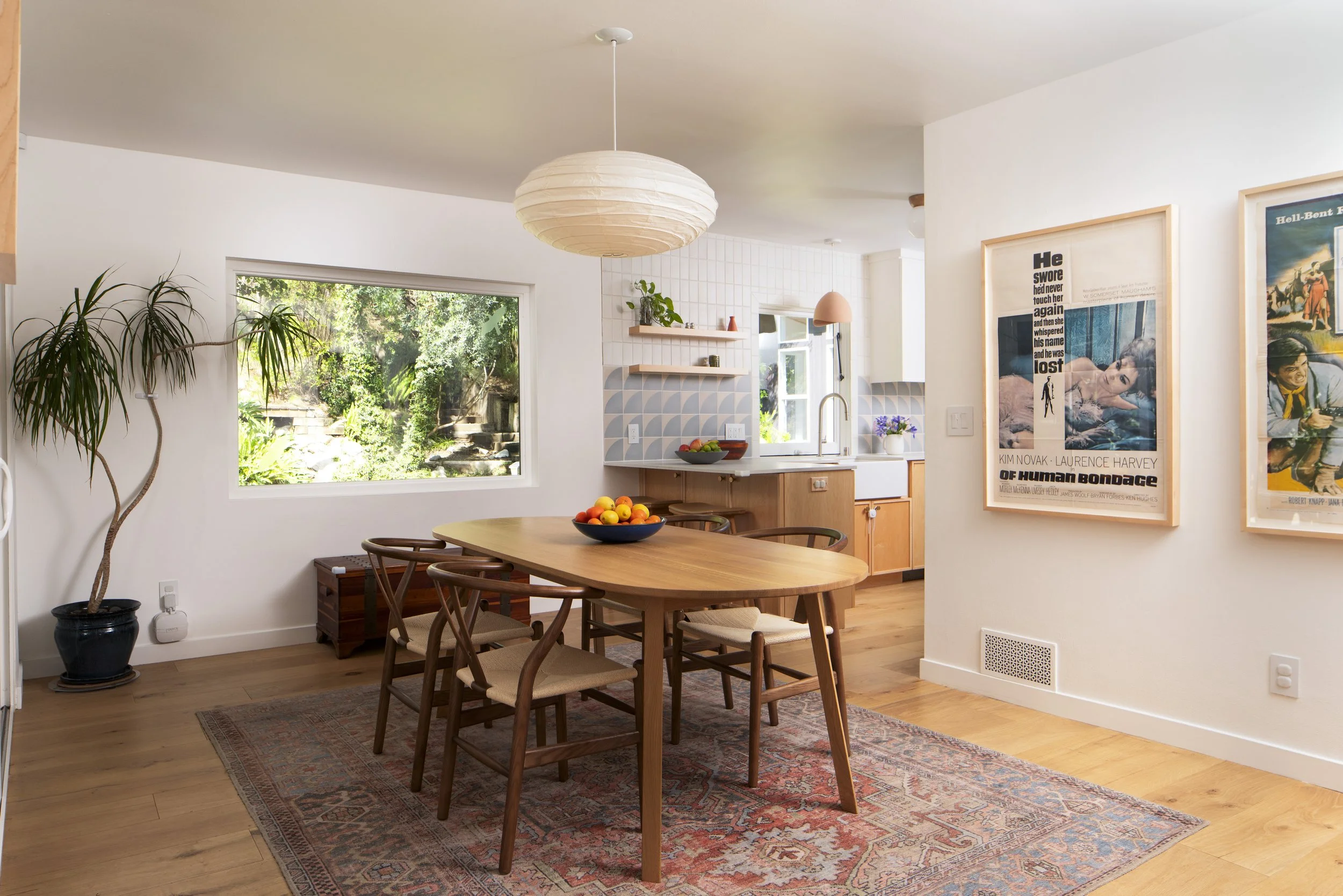 Bright dining area with wooden table, six matching chairs, a bowl of fruit, large window showing greenery, and modern art posters on the wall.