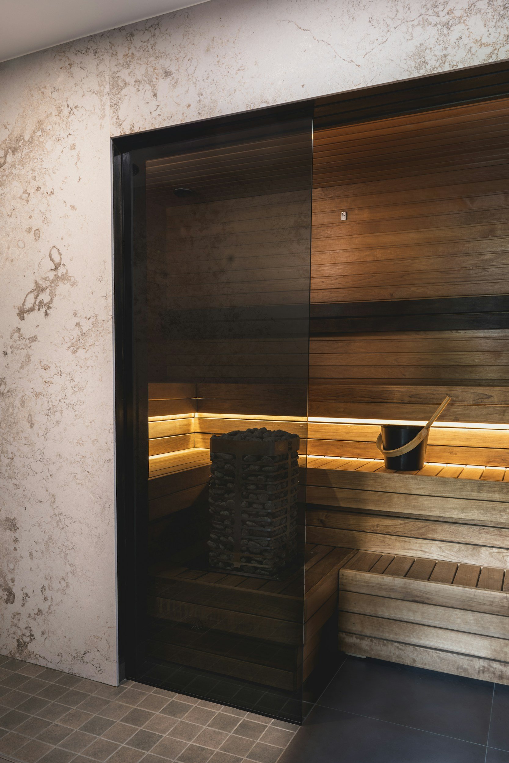 Interior of a sauna with wooden benches, a bucket with a wooden ladle, a heater with stones, and a glass door.