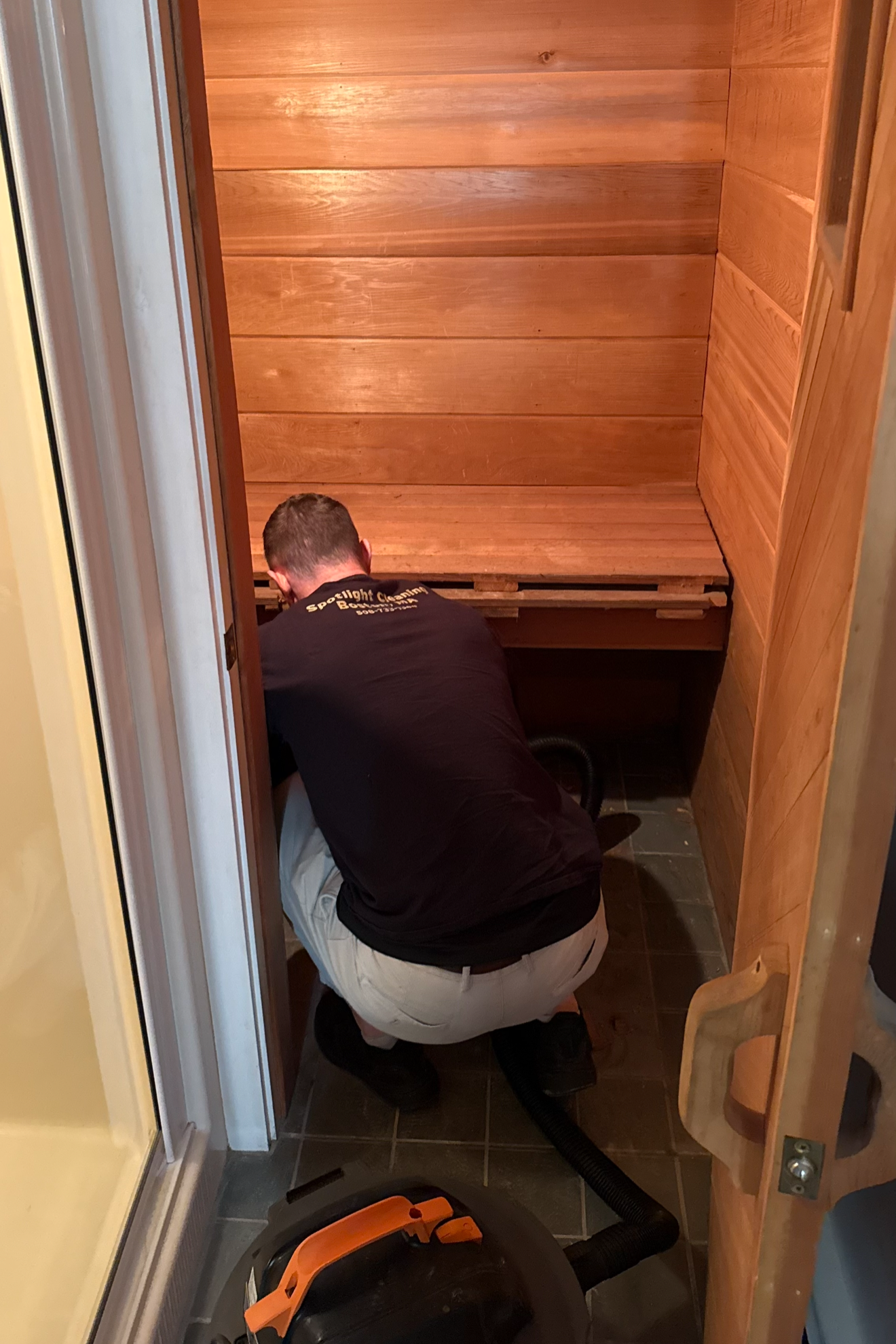 A man kneeling inside a small wooden closet with a vacuum hose, cleaning or inspecting the area.