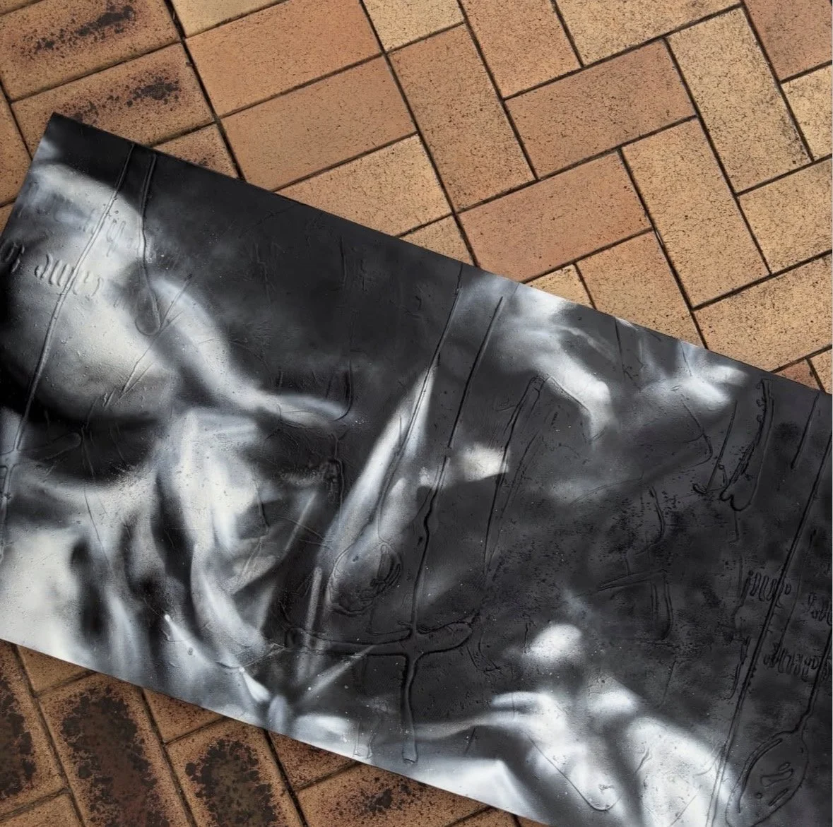 Close-up of a black and white spray-painted mural or artwork on a metal sheet, resting on a brick floor.
