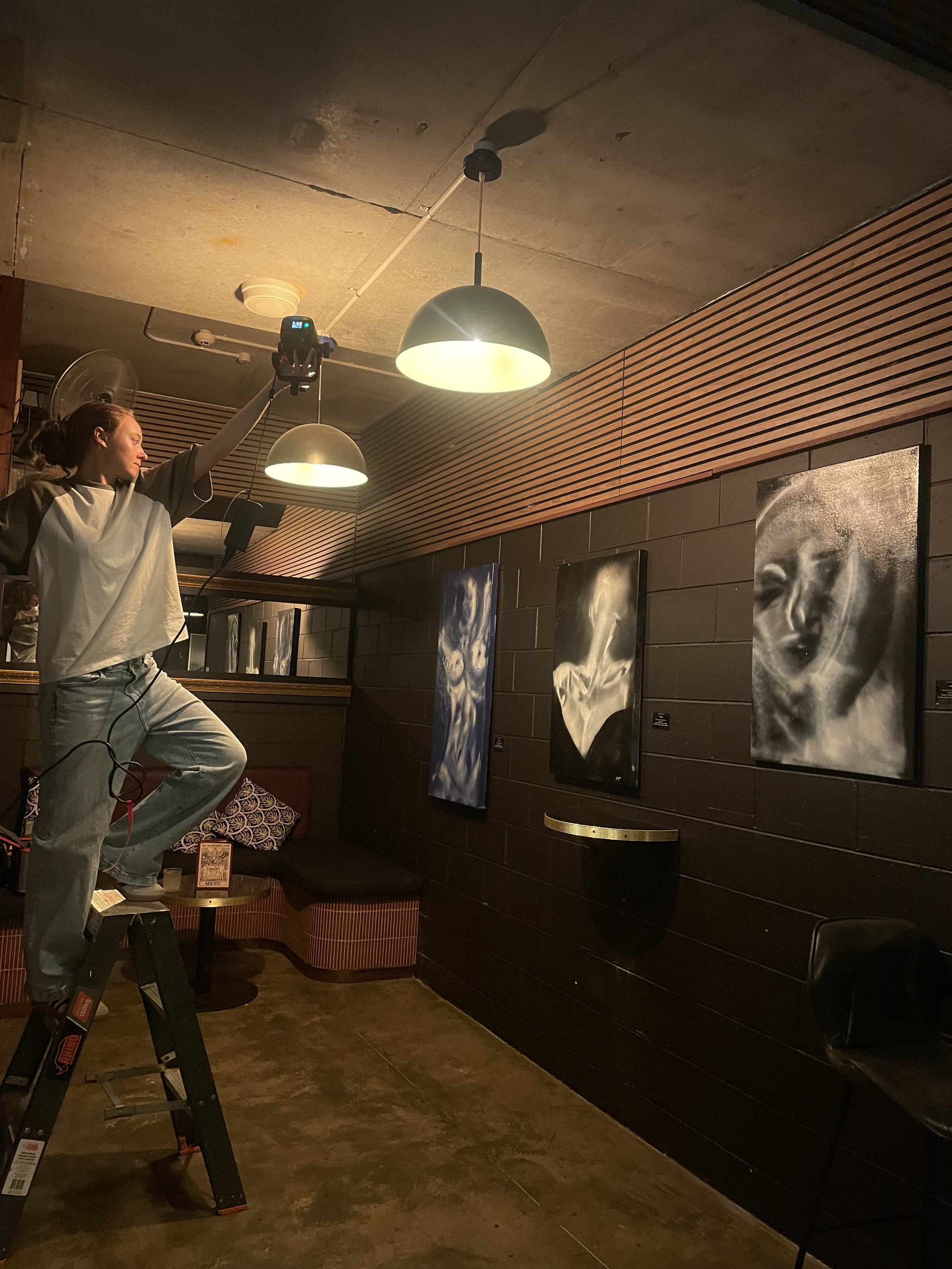 A person standing on a ladder adjusting lighting in an art gallery with abstract black and white paintings on the wall.