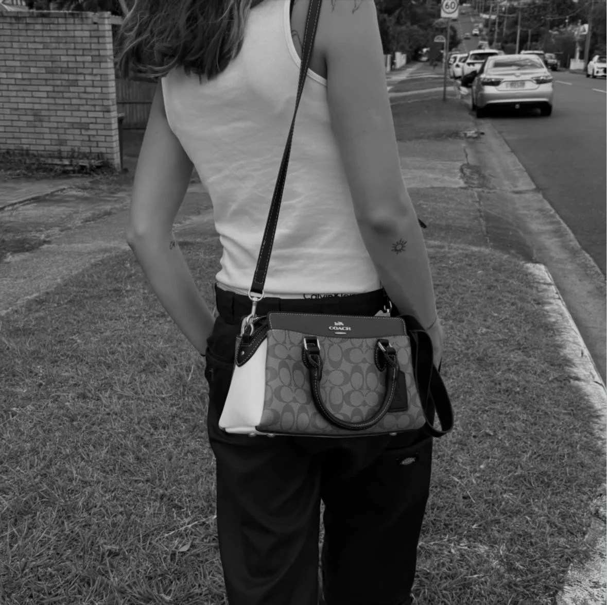 A woman stands on a sidewalk with her back to the camera, wearing a sleeveless top, dark pants, and carrying a Coach handbag with a patterned design and a shoulder strap. She has a temporary tattoo of a sun on her left arm.