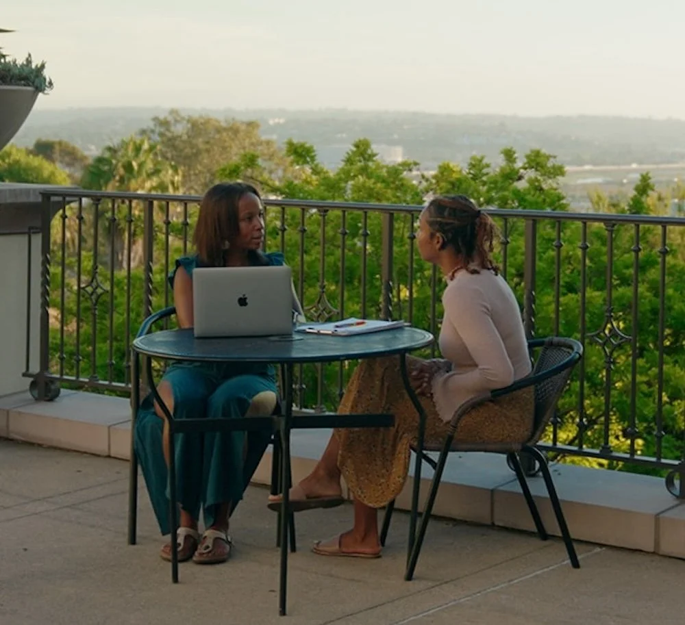 Two brown-skinned female professionals having a coaching conversation at an outdoor table with a laptop.