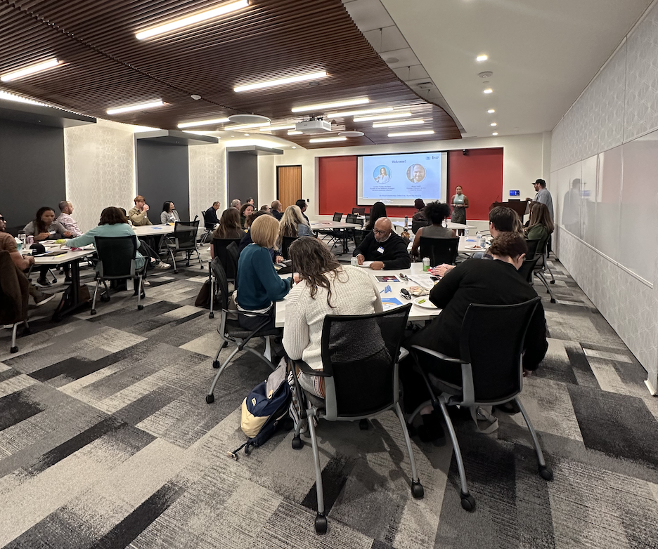 People seated at round tables in a conference room while one presenter stands at the front near a large screen.