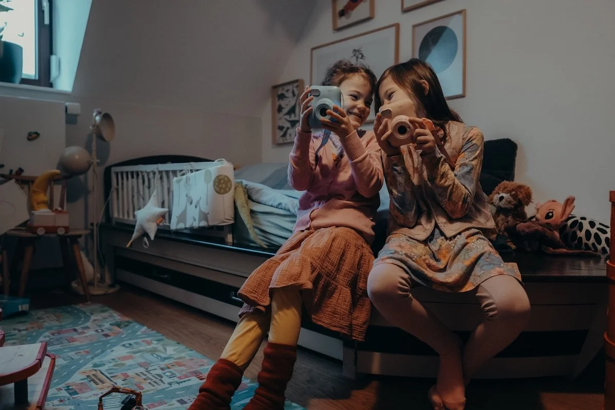 Zwei Schwestern sitzen nebeneinander auf einem Bett im Kinderzimmer, während sie die Köpfe aneinander halten und sich anlachen. Dabei haben beide eine Sofortbildkamera in der Hand.