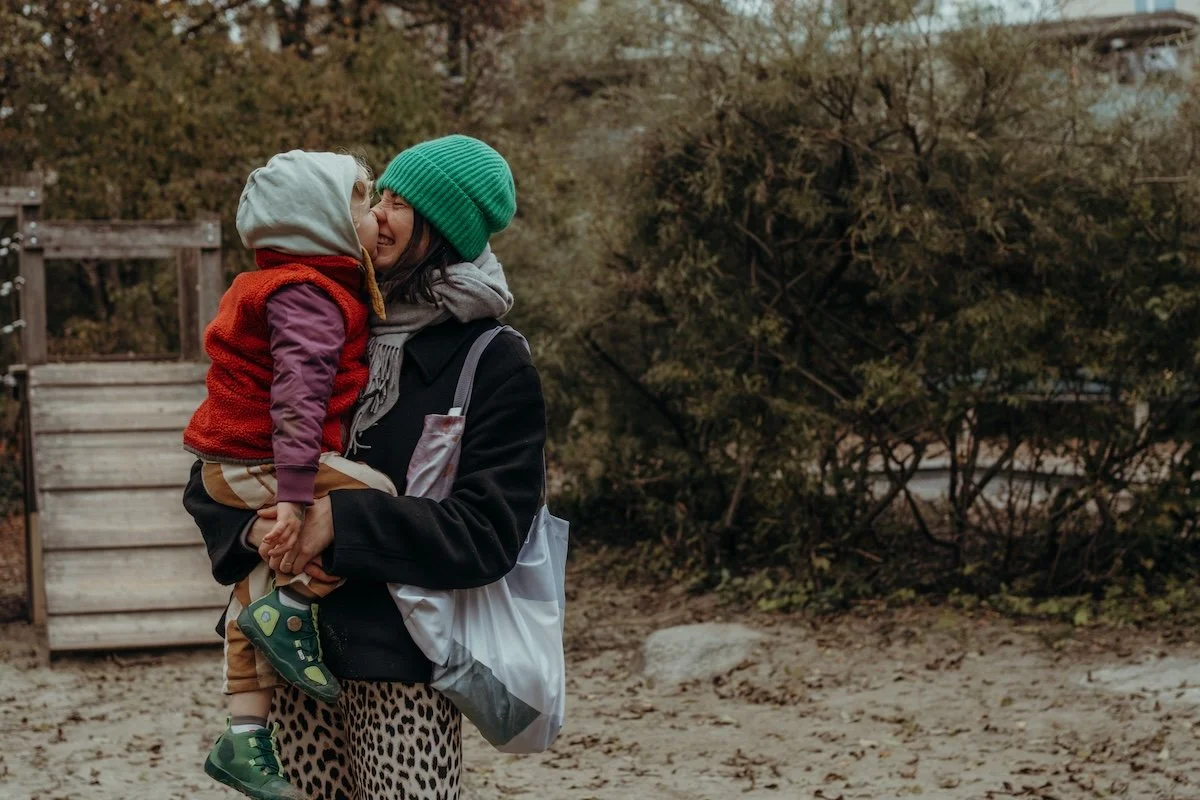 Eine Mama gibt ihrem Sohn einen Nasenkuss auf einem Spielplatz, beide lachen dabei.