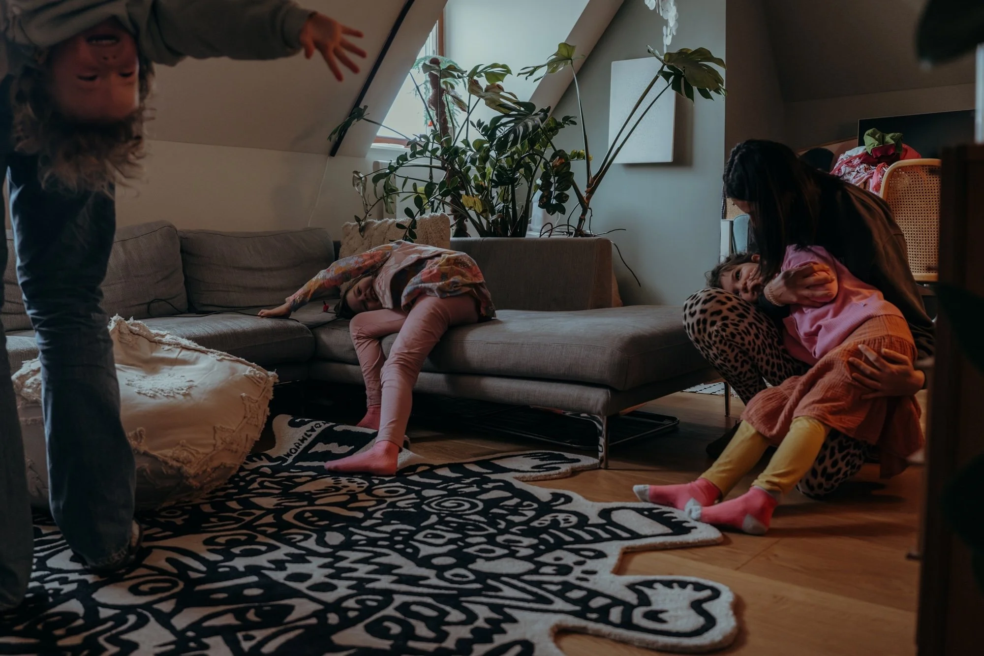 Eine Familie mit drei Kindern ist in ihrem Wohnzimmer, wobei das jüngste Kind kopfüber vom Papa an den Beinen gehalten wird. Die älteste Tochter liegt auf dem Sofa, während die andere Tochter in Mamas Armen liegt und liebevoll getröstet wird.