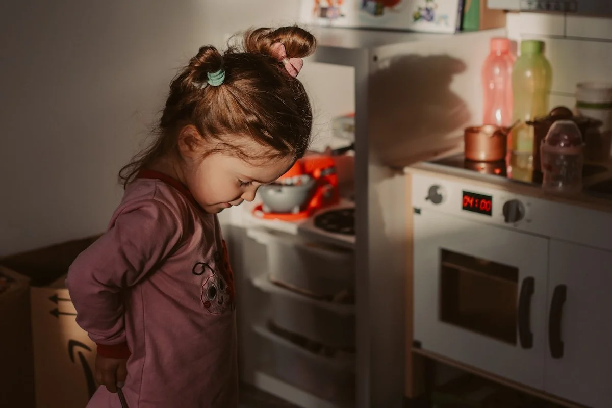 Ein Mädchen mit zwei Zöpfen steht im Schlafanzug an einer Spielküche und wird von der Morgensonne angestrahlt.