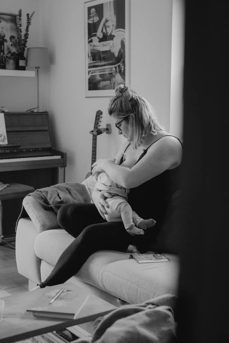 Eine Mama stillt ihr neugeborenes Baby im Wohnzimmer und sitzt dabei auf dem Sofa.