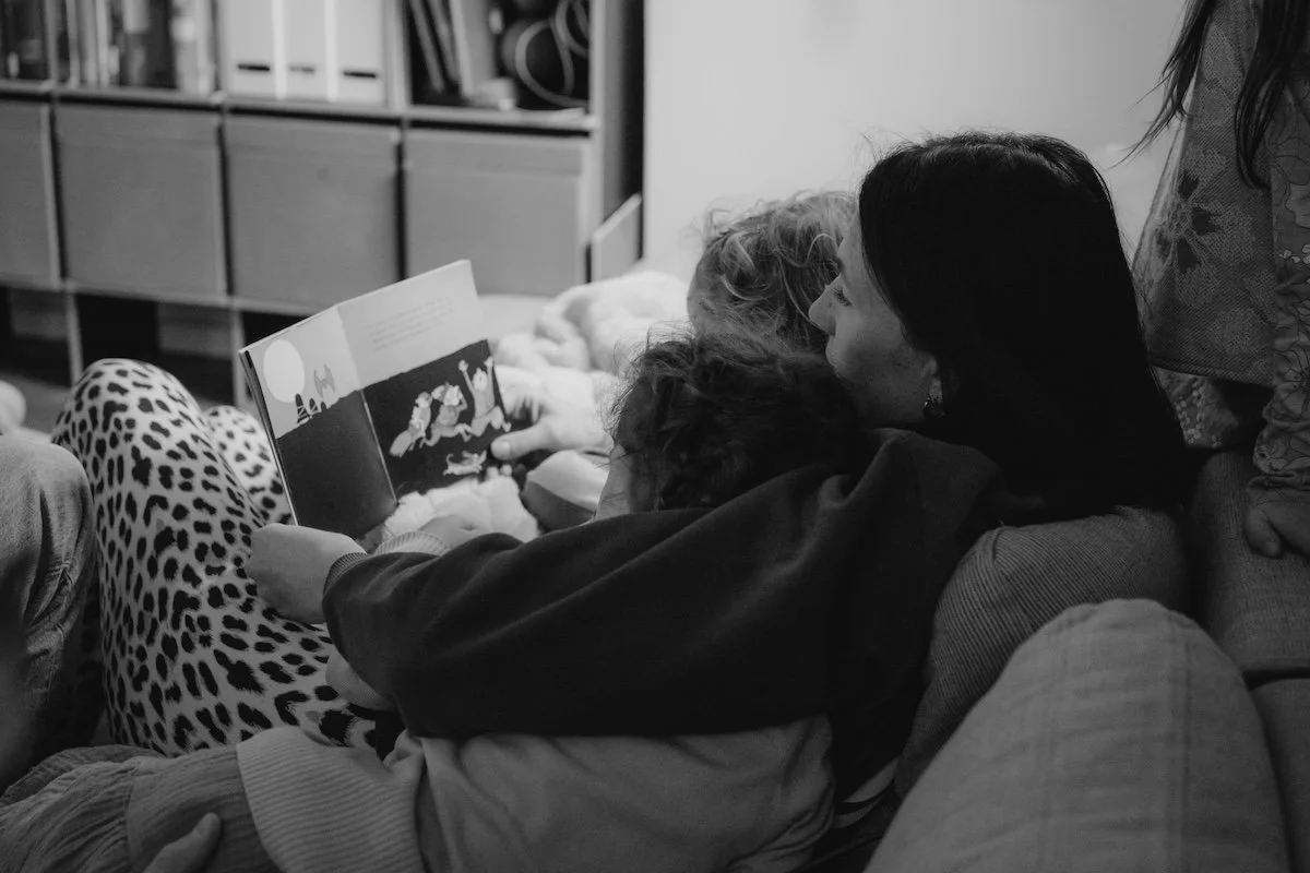 Eine Mama liegt mit ihren zwei Kindern im Arm auf dem Sofa und liest ihnen ein Buch vor.
