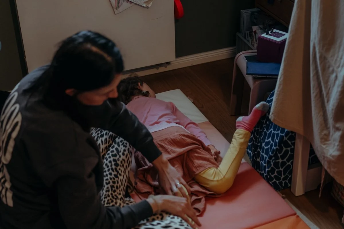 Eine Mama massiert ihre Tochter, die im Kinderzimmer auf einer Matte auf dem Boden liegt.