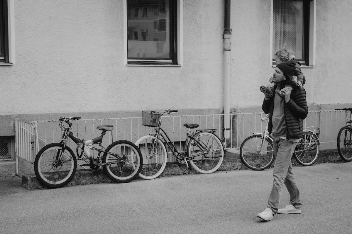 Der Papa trägt seinen Sohn auf den Schultern und läuft an einer Hauswand entlang, an welcher viele Fahrräder stehen.