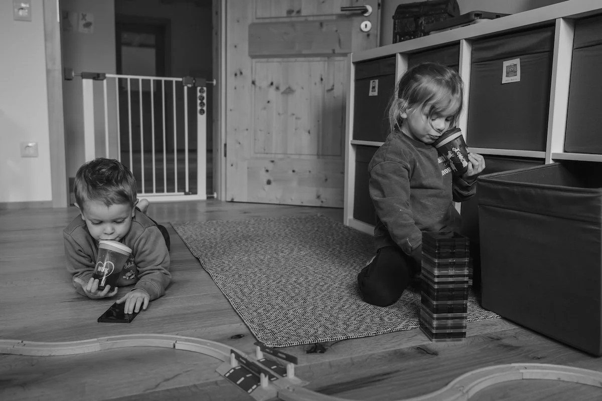 Zwei Geschwister spielen im Kinderzimmer mit einer Holzeisenbahn und Magnetbausteinen, während beide aus einem Becher mit Strohhalm trinken.