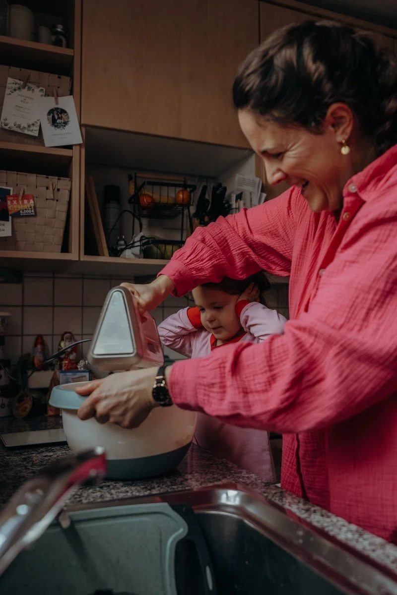 Eine Mama backt mit ihrer Tochter, wobei die Mama das Rührgerät in der Hand hält und lacht und sich die Tochter die Ohren zuhält.
