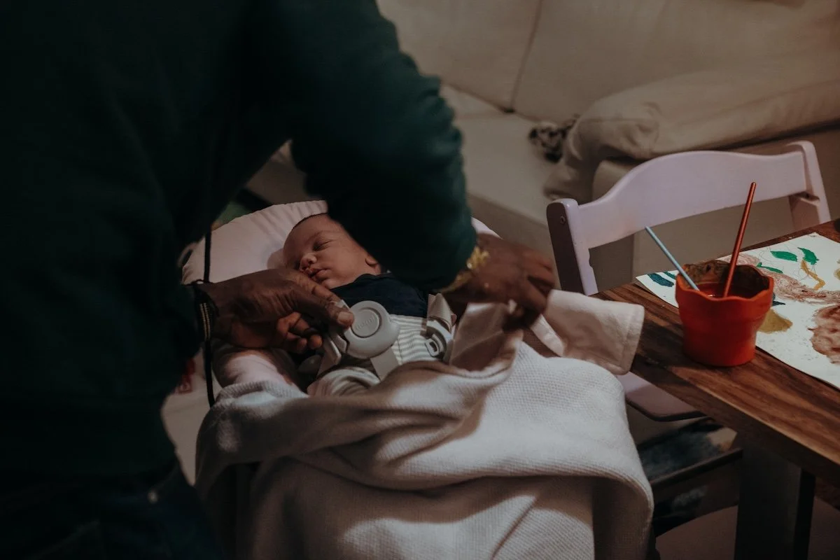 Der Papa schnallt seine neugeborene Tochter sein Baby ab, welches zugedeckt und schlafend in einem Neugeborenenaufsatz eines Hochstuhls liegt.