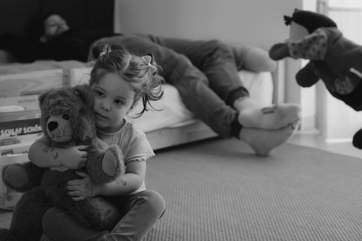 Ein junges Mädchen hält einen großen Kuschelbären im Arm, während ihr Papa im Hintergrund auf dem Kinderbett liegt und die Mama eine Handpuppe zu ihrer Tochter hält.