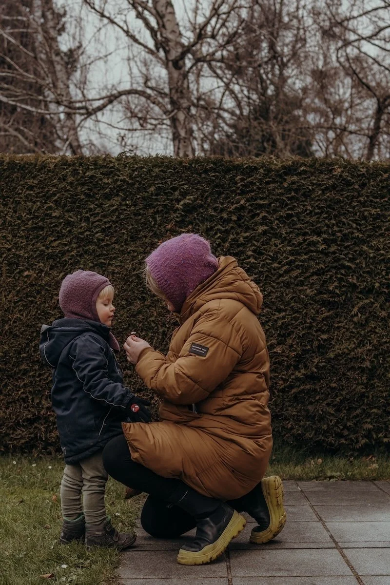Eine Mama kniet mit einer kleinen Blumen in der Hand vor ihrem kleinen Sohn, beide haben eine lila Schlupfmütze an.