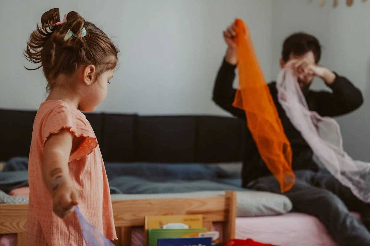 Ein Mädchen mit Tattooresten auf dem Arm und zwei Zöpfen steht mit Seidentüchern in der Hand im Kinderzimmer, während der Papa im Hintergrund ebenfalls mit Tüchern spielt.