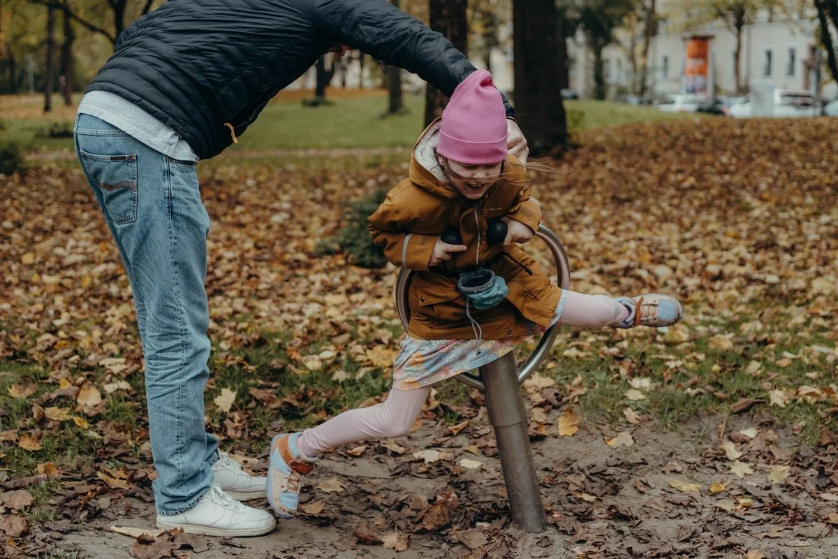 Der Papa schubst seine Tochter an, welche auf einem drehenden Spielgerät auf einem Spielplatz sitzt. Das Mädchen lacht dabei.